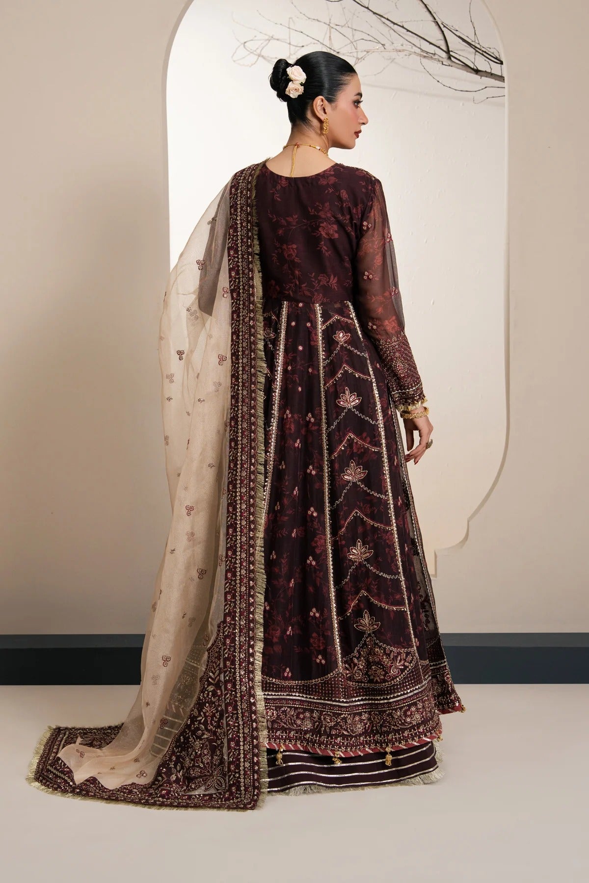 A woman modeling a traditional dark maroon anarkali suit with gold embroidery and a beige printed dupatta, shown from the back in a studio setting.