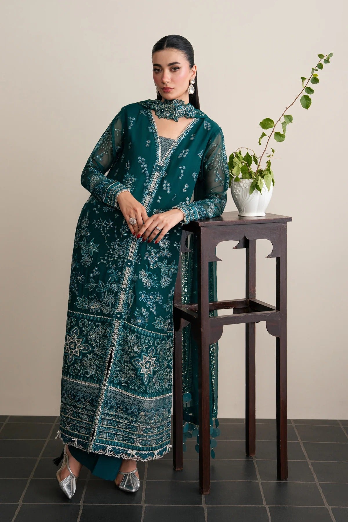 A woman models a traditional long teal green embroidered kurta set with a matching dupatta, standing next to a dark wooden table with a potted plant against a beige wall.