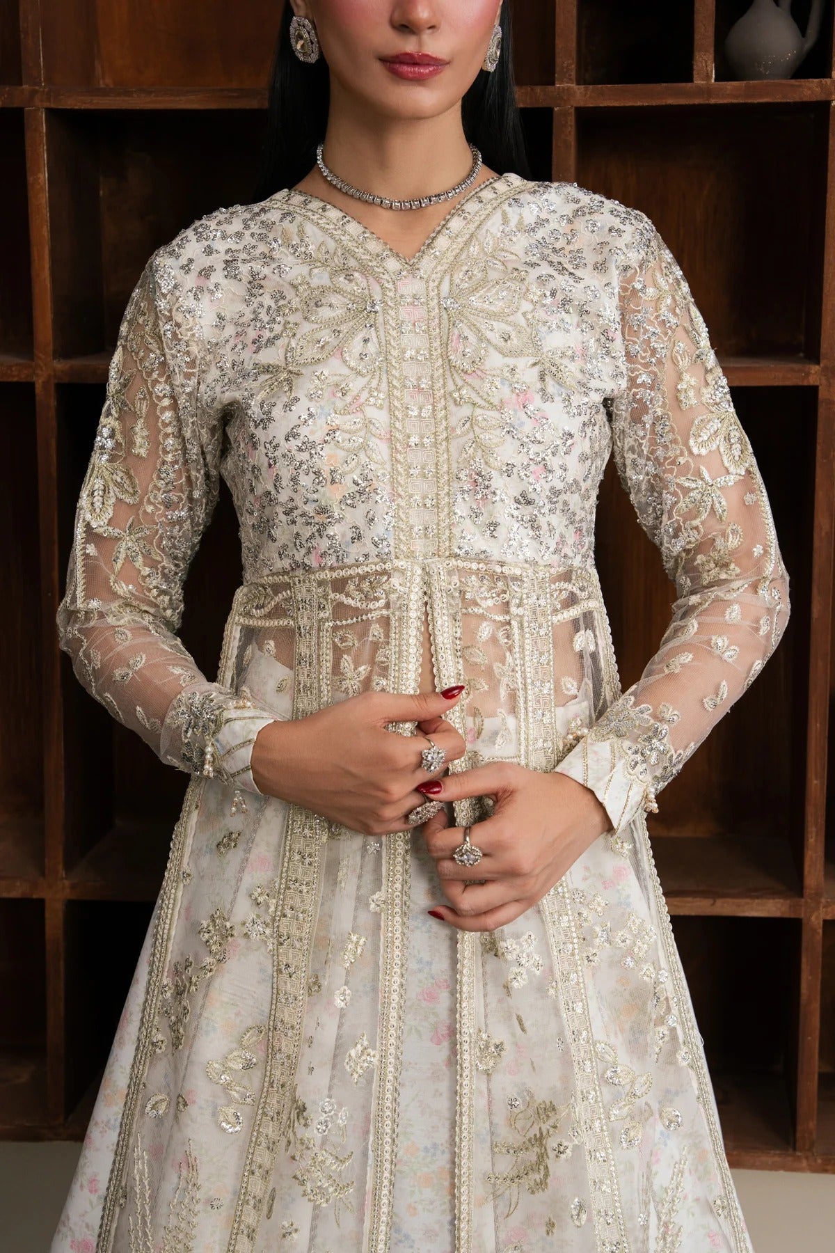 A woman wearing a long-sleeved cream-colored embellished dress with intricate gold and silver embroidery, standing in front of a dark wooden shelving unit.