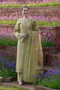Woman in a green embroidered traditional outfit standing in a garden with flowers and brick steps.