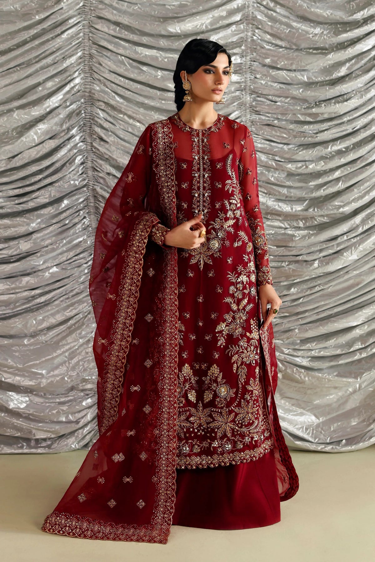 A woman modeling a deep red traditional Indian outfit with intricate gold embroidery and a matching sheer dupatta, posed against a silver draped backdrop.