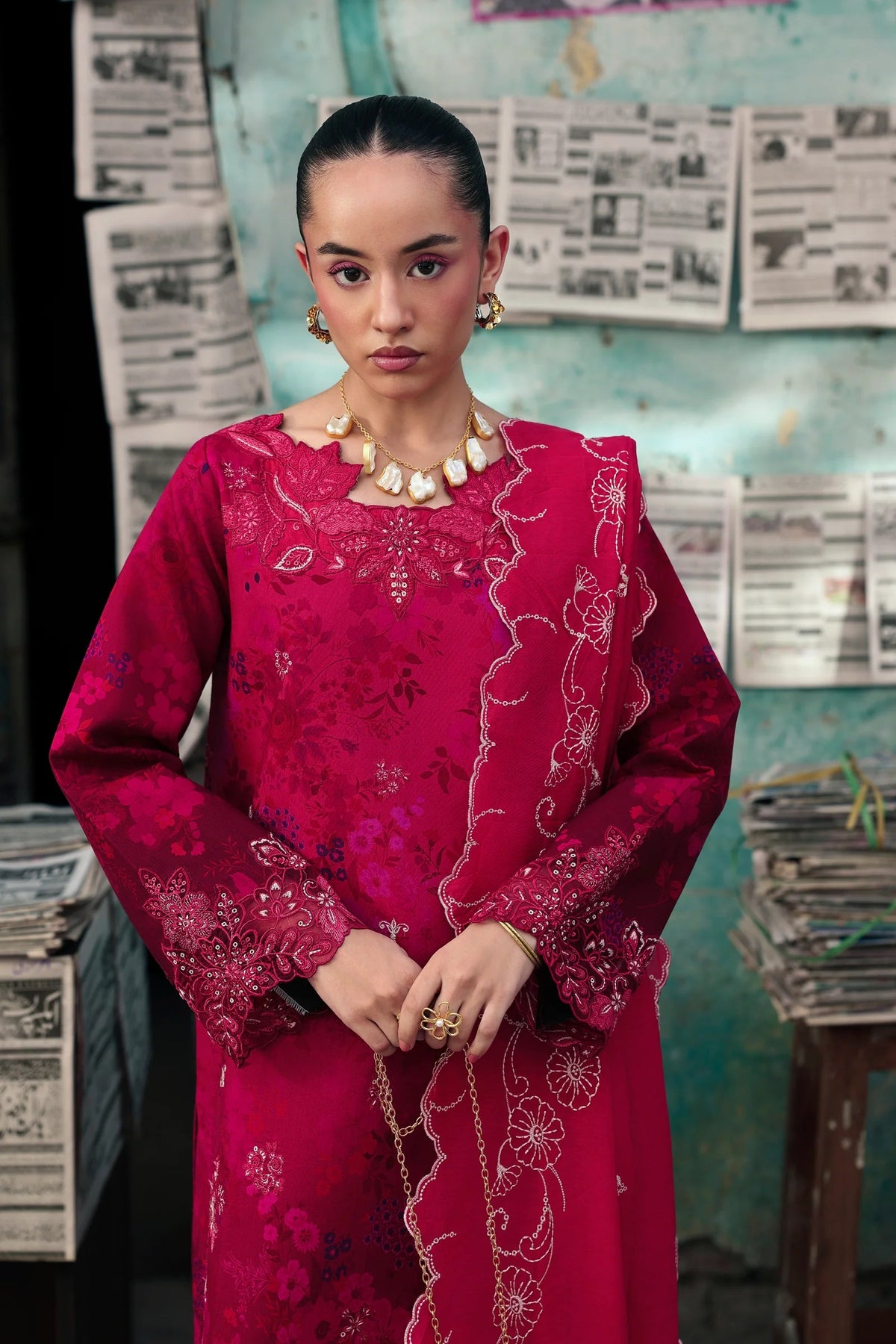 A woman wearing a vibrant magenta-pink traditional embroidered dress with gold detailing, standing in front of a rustic wall covered in newspaper clippings.
