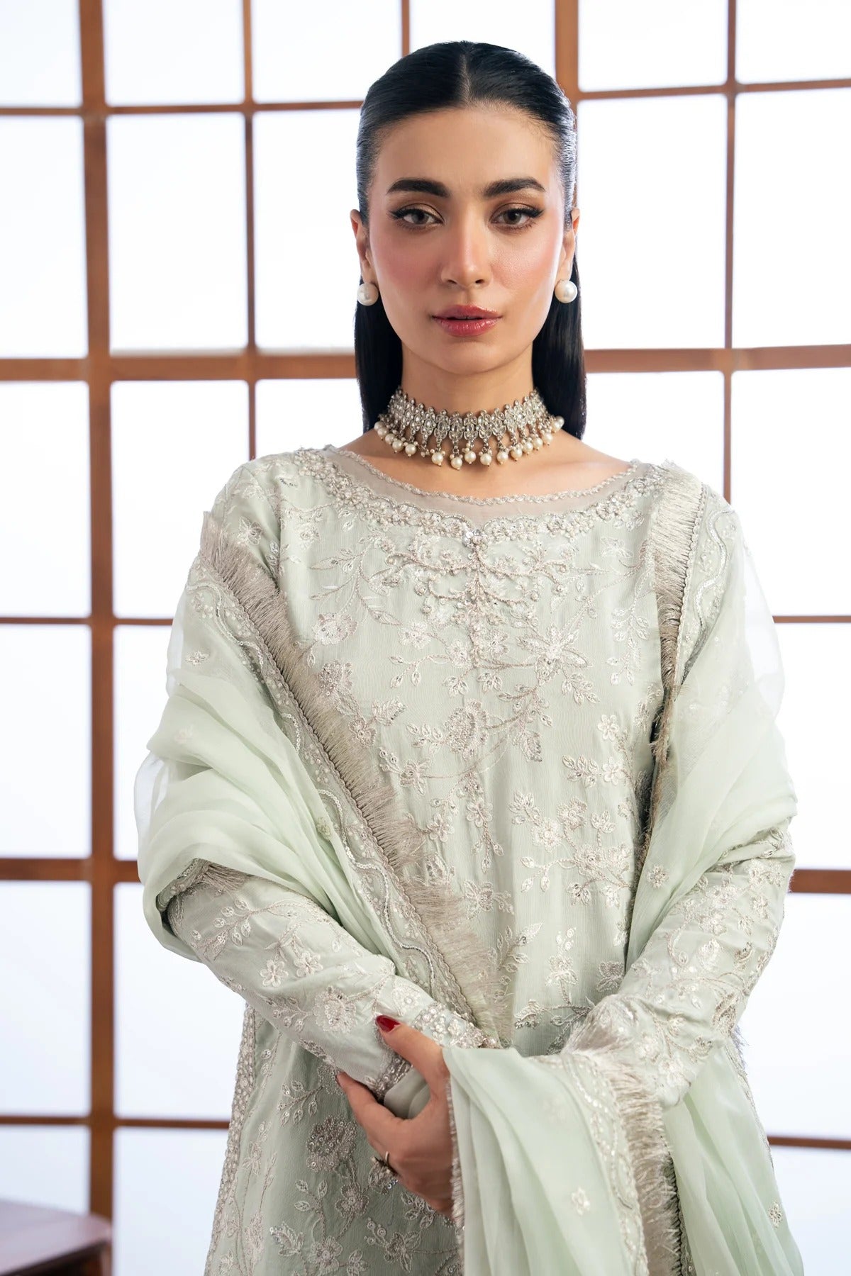 A woman modeling a light sage green embroidered traditional outfit with a matching dupatta, set against a white background with a wooden grid screen.