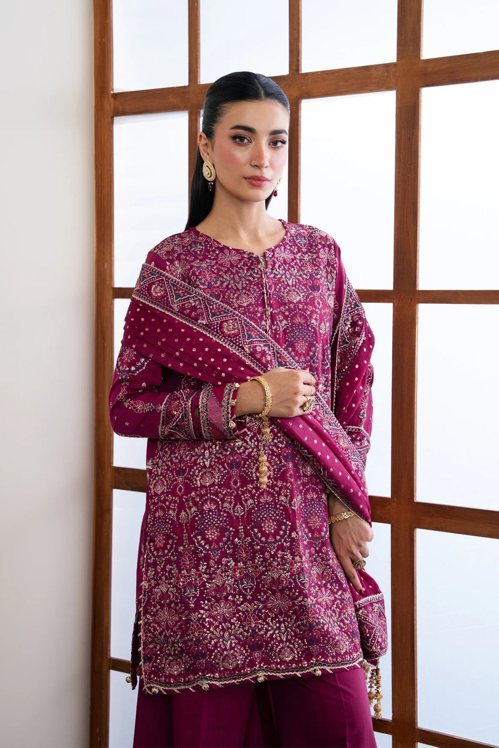 A woman models a traditional three-piece salwar kameez suit in deep magenta with intricate gold embroidery and a matching patterned dupatta, standing in front of a wooden grid window.
