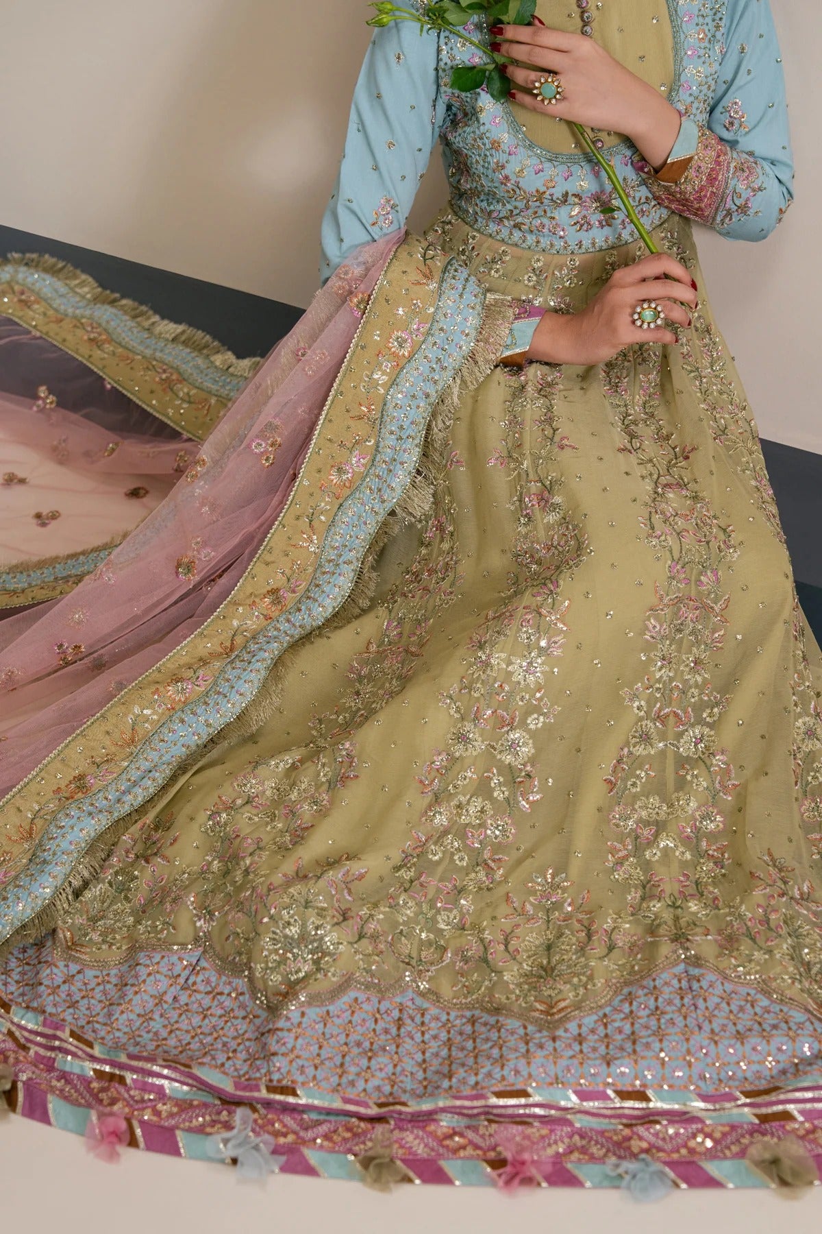 A close-up of a model wearing an ornate traditional South Asian outfit, featuring a light blue long-sleeve top paired with a heavily embroidered olive green skirt and a pink dupatta with gold borders.