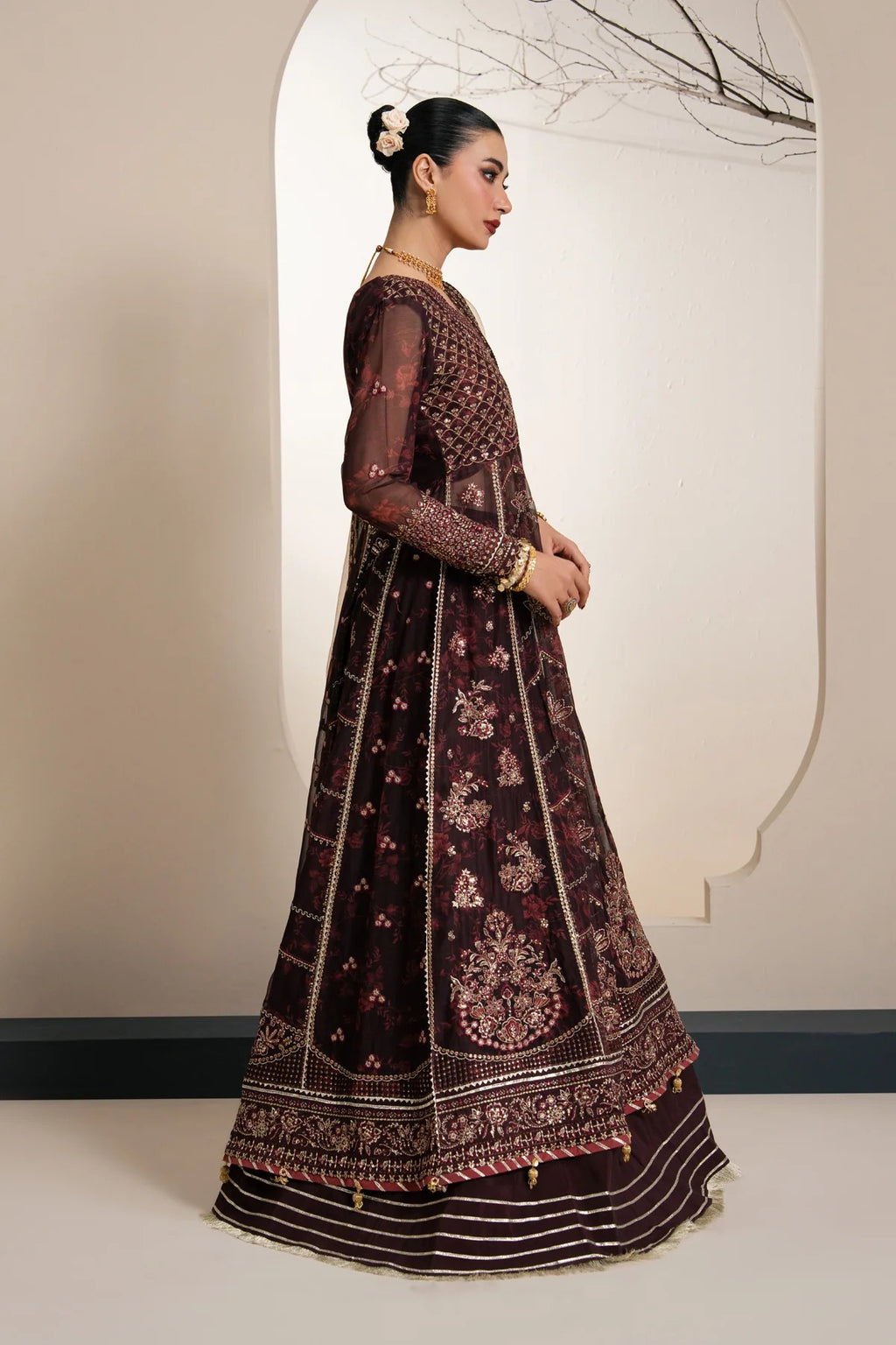 A woman modeling a dark brown embroidered Anarkali gown with intricate gold and pink floral patterns, standing in profile against a beige wall with a decorative arch.