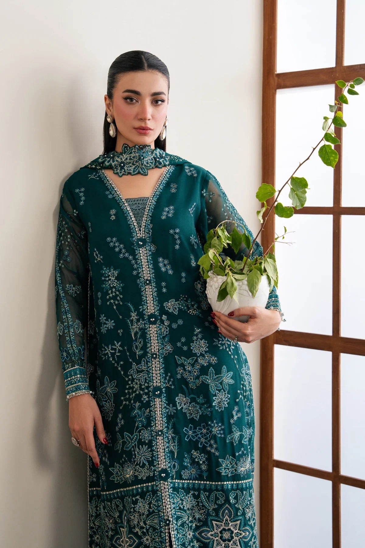 A woman models a dark teal green ethnic kurta with intricate white and silver floral embroidery, holding a potted green plant in front of a white wall and wooden window frame.