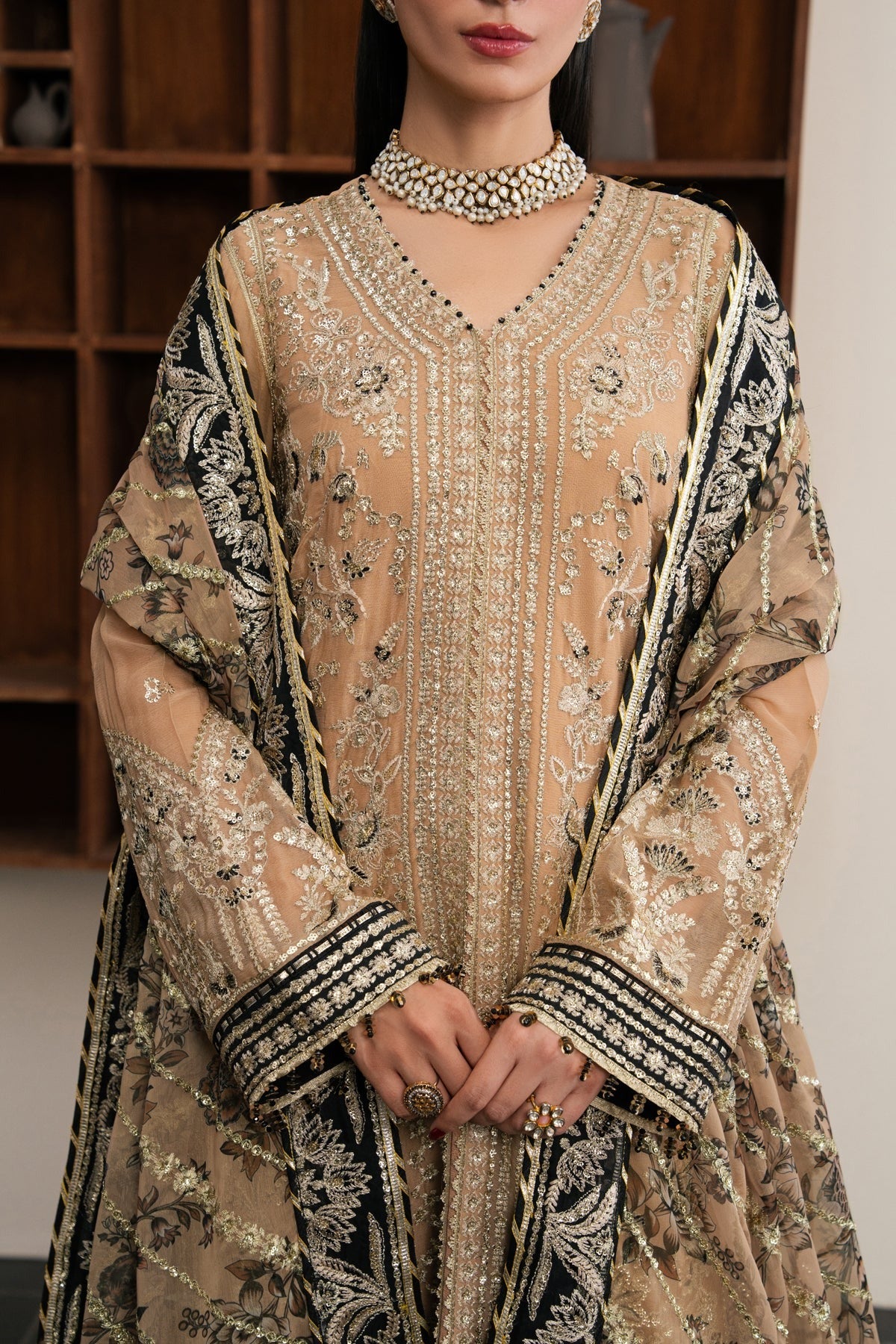 A woman wearing an ornate beige and black embroidered traditional outfit with detailed metallic embellishments, a matching sheer dupatta, and a pearl choker, standing indoors in front of a wooden shelving unit.