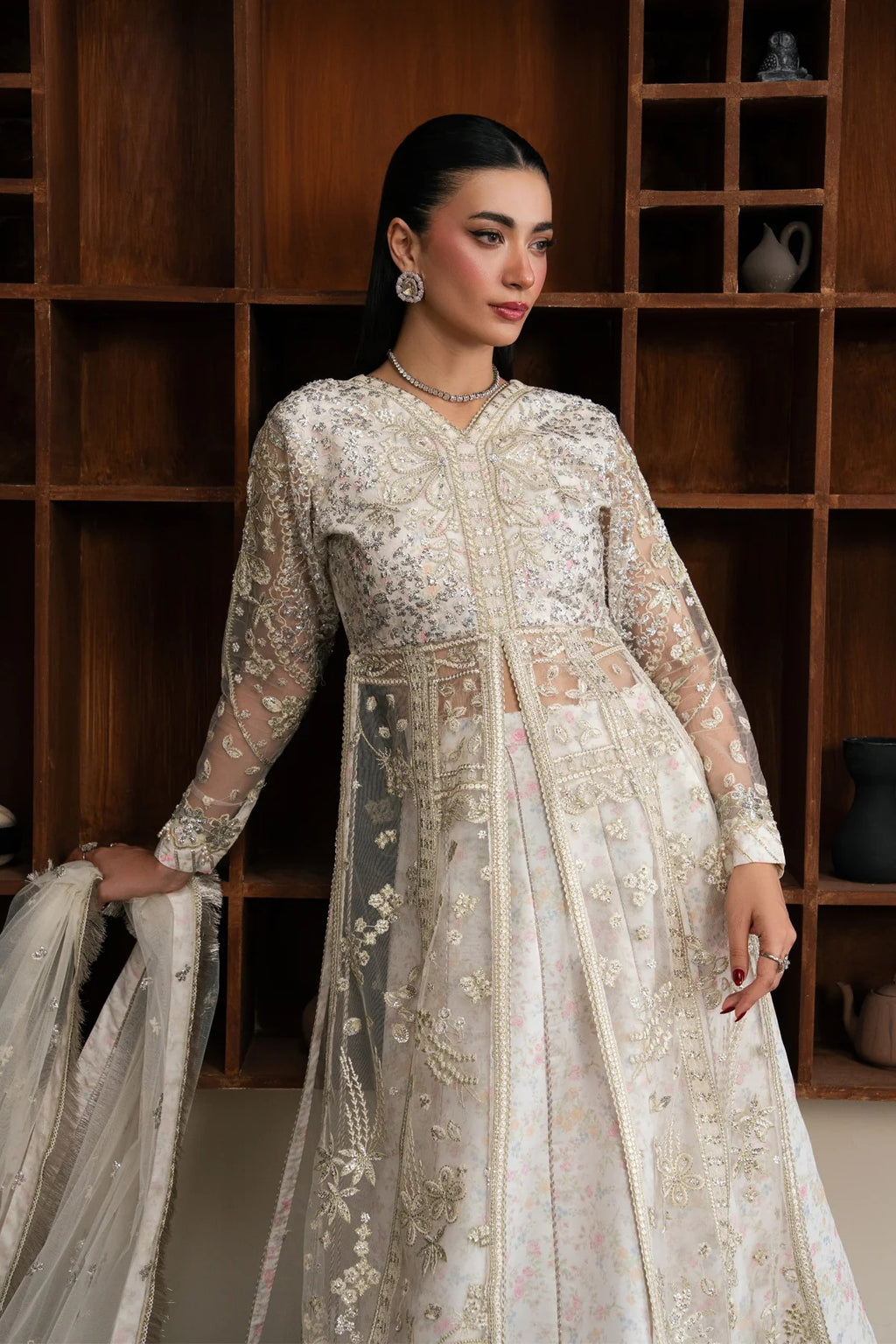 A woman models a heavily embroidered off-white and beige traditional gown with sheer sleeves and a matching dupatta, posed in front of a dark wooden shelving unit.