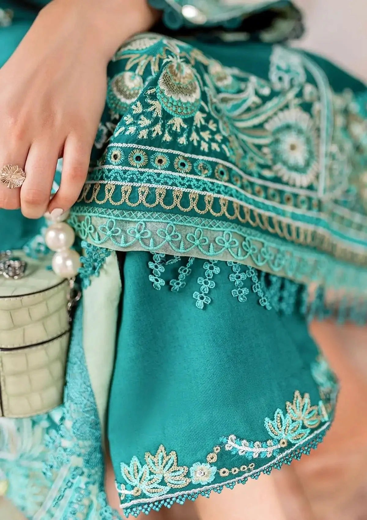 A close-up of a teal embroidered traditional garment with intricate floral and geometric patterns, held by a hand wearing a ring next to a pearl-trimmed clutch bag.