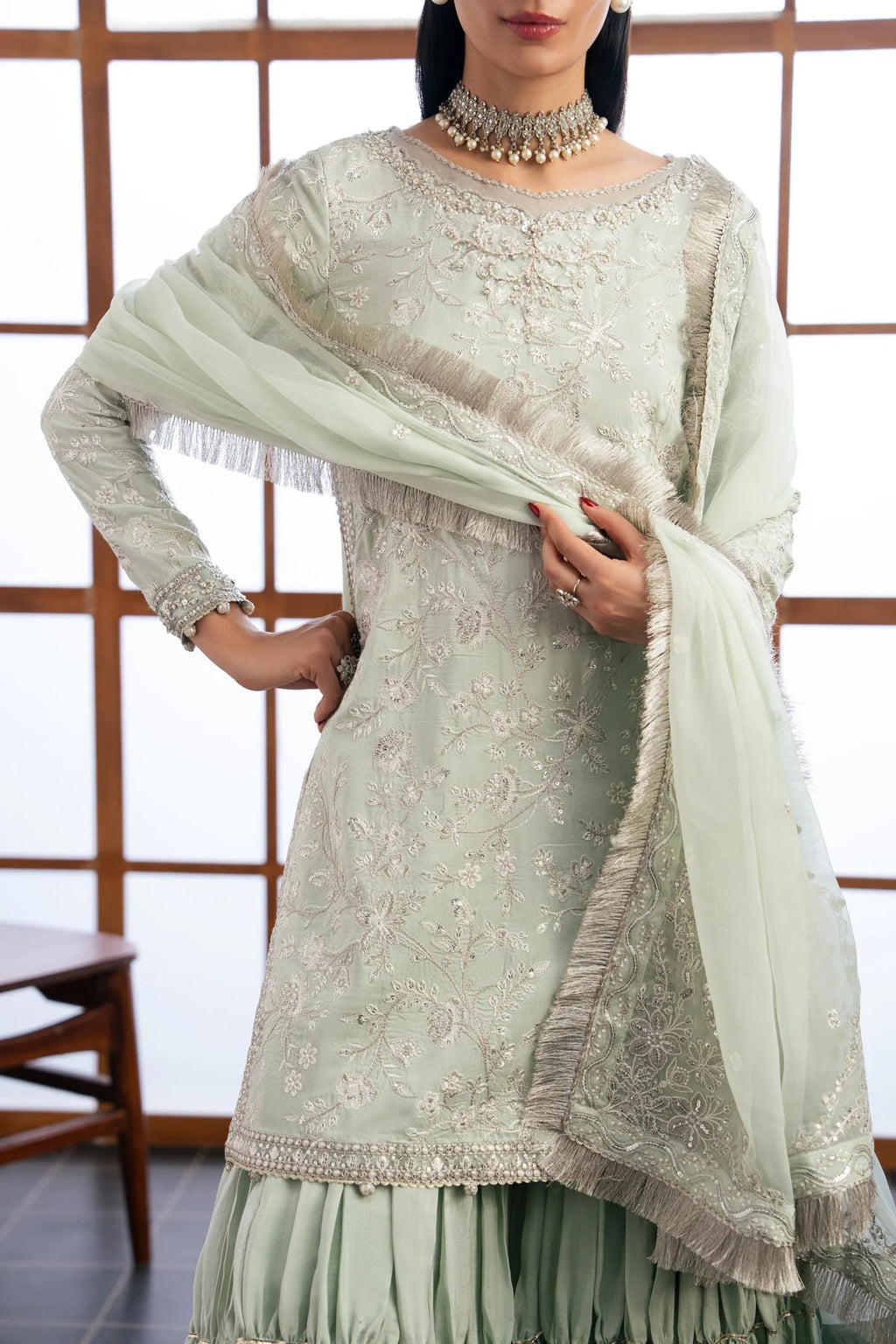 A woman models a light sage green traditional Indian outfit with intricate silver embroidery, a matching pleated skirt, and a sheer dupatta with silver fringe, set against a background of wooden-framed windows.