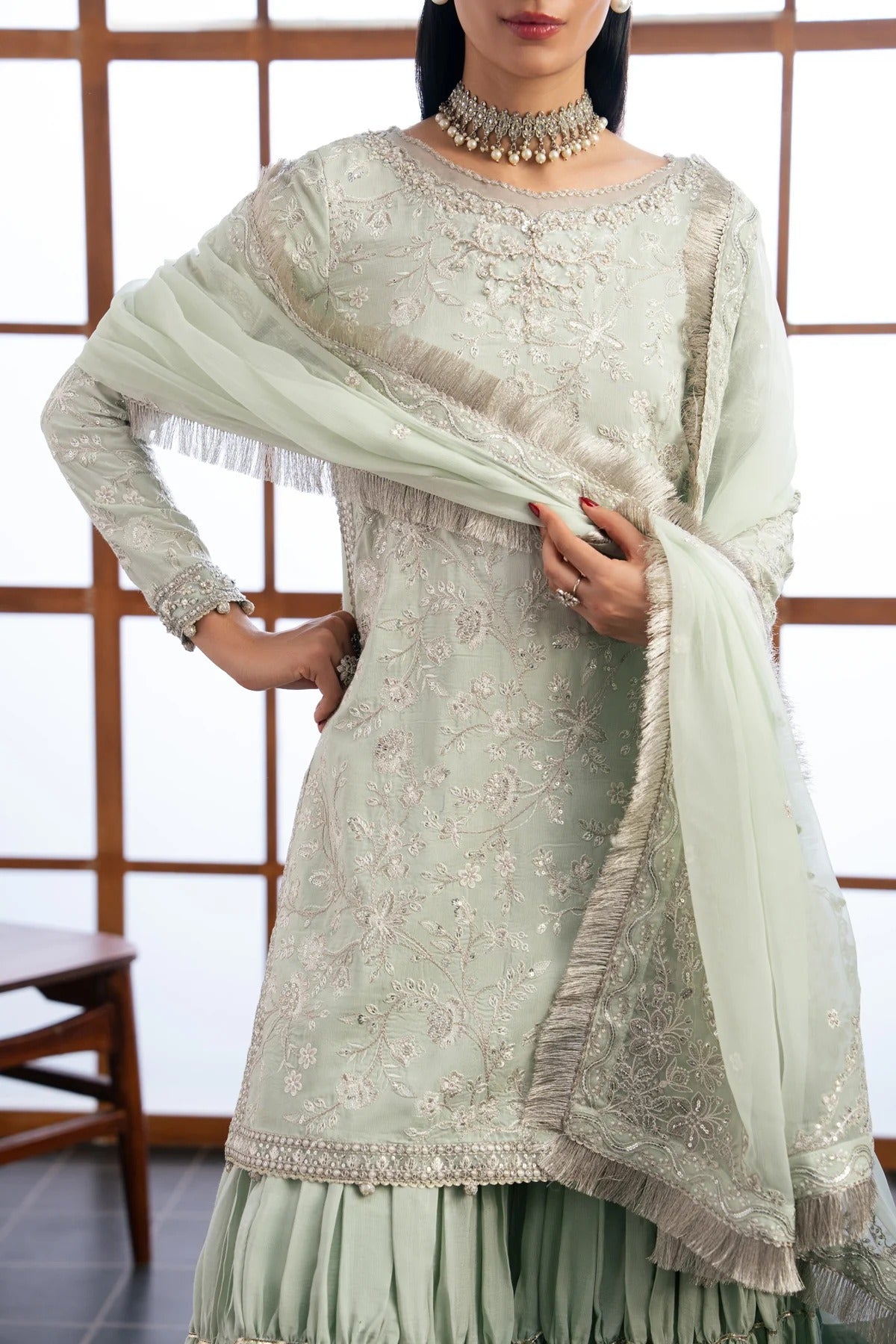 A woman models a light sage green traditional Indian outfit with intricate silver embroidery, a matching pleated skirt, and a sheer dupatta with silver fringe, set against a background of wooden-framed windows.