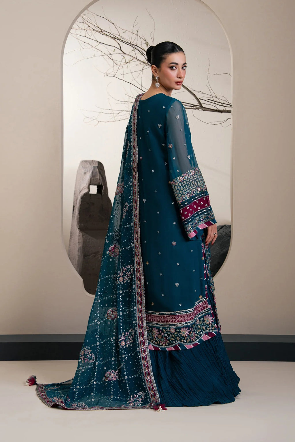 A woman modeling a traditional South Asian outfit, shown from the back. The outfit consists of a deep teal blue embroidered kurta with matching flared pants and a sheer embroidered dupatta.