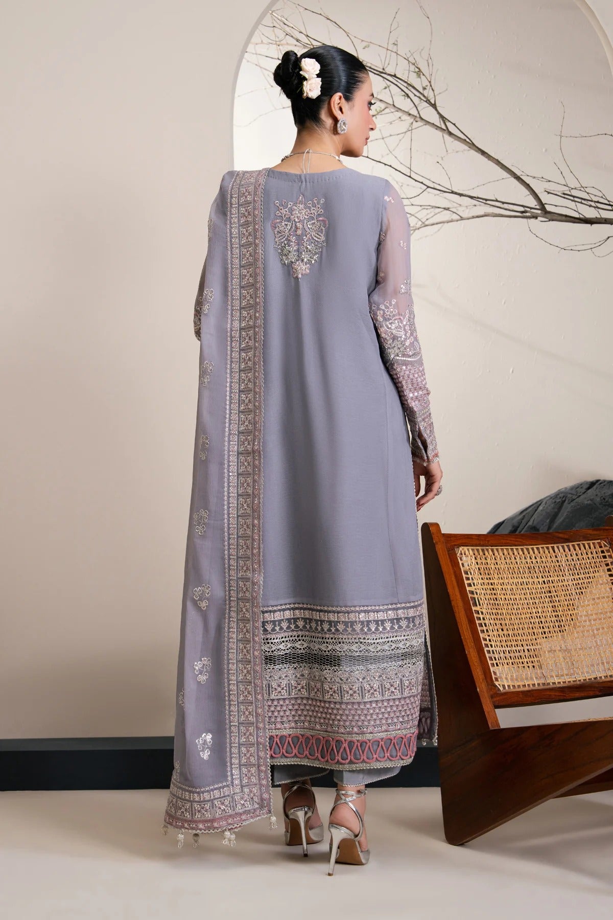 A woman stands with her back to the camera wearing a traditional long lavender-gray salwar kameez suit with intricate multicolor embroidery and a matching dupatta. She is posed in a styled indoor setting with a wooden chair and neutral wall background.