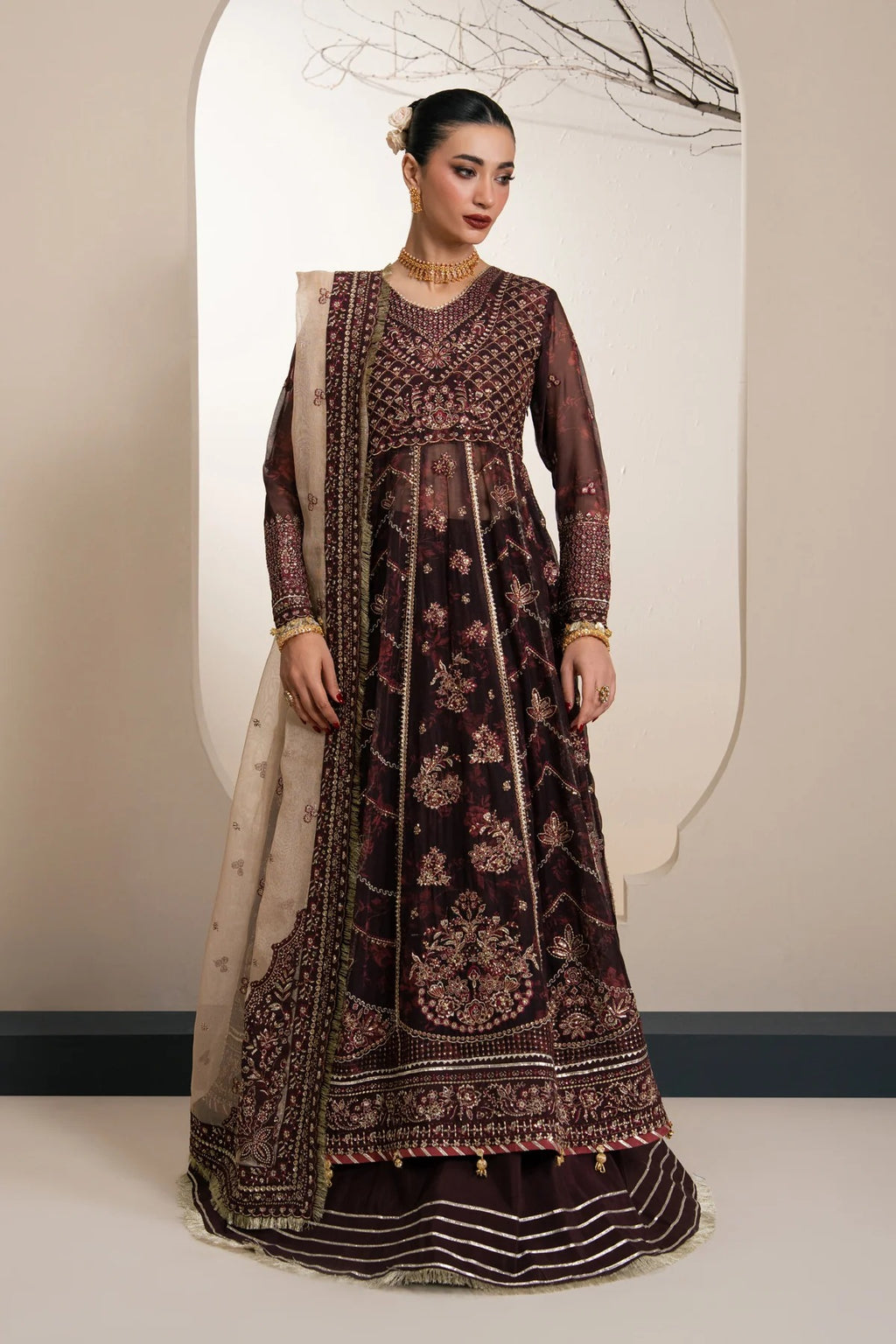 A woman models a traditional Indian outfit consisting of a dark plum velvet anarkali-style dress with intricate gold embroidery, a matching embroidered dupatta, and a striped skirt, set against a minimalist beige studio background.