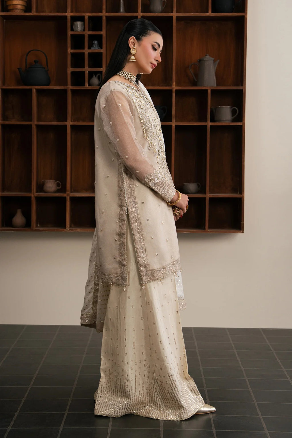A woman models a traditional beige and gold embroidered ethnic outfit with a sheer dupatta, standing in front of a dark wooden shelving unit.