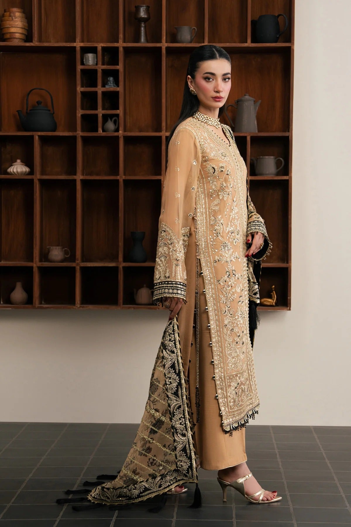 A woman models a traditional beige and gold embroidered kurta set with a matching patterned dupatta, standing in front of a wooden display shelf.