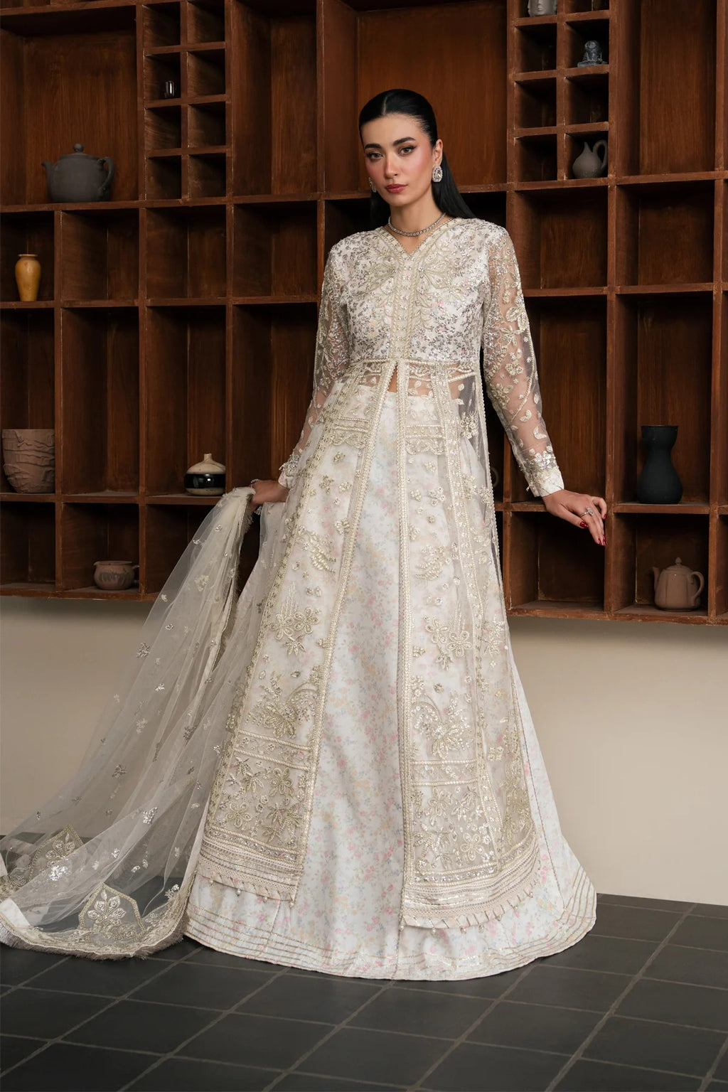 A woman models a traditional Indian anarkali gown in an off-white/cream color with intricate gold embroidery, paired with a matching sheer dupatta, set against a dark wood shelving background.