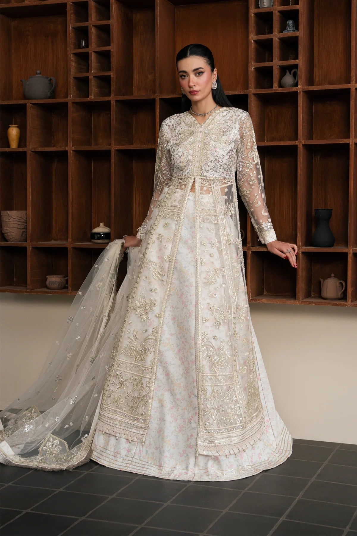 A woman models a traditional Indian anarkali gown in an off-white/cream color with intricate gold embroidery, paired with a matching sheer dupatta, set against a dark wood shelving background.