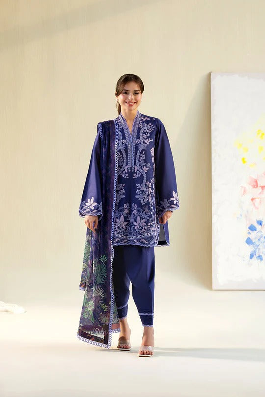 Woman wearing a blue embroidered outfit standing in a minimalistic room with a painting on the wall.