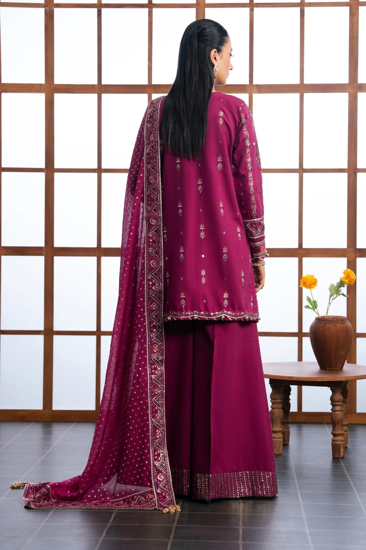A woman wearing a deep magenta three-piece traditional outfit with gold embroidery, shown from the back. The set includes a long kurta, wide-leg pants, and a matching dupatta draped over her shoulder.