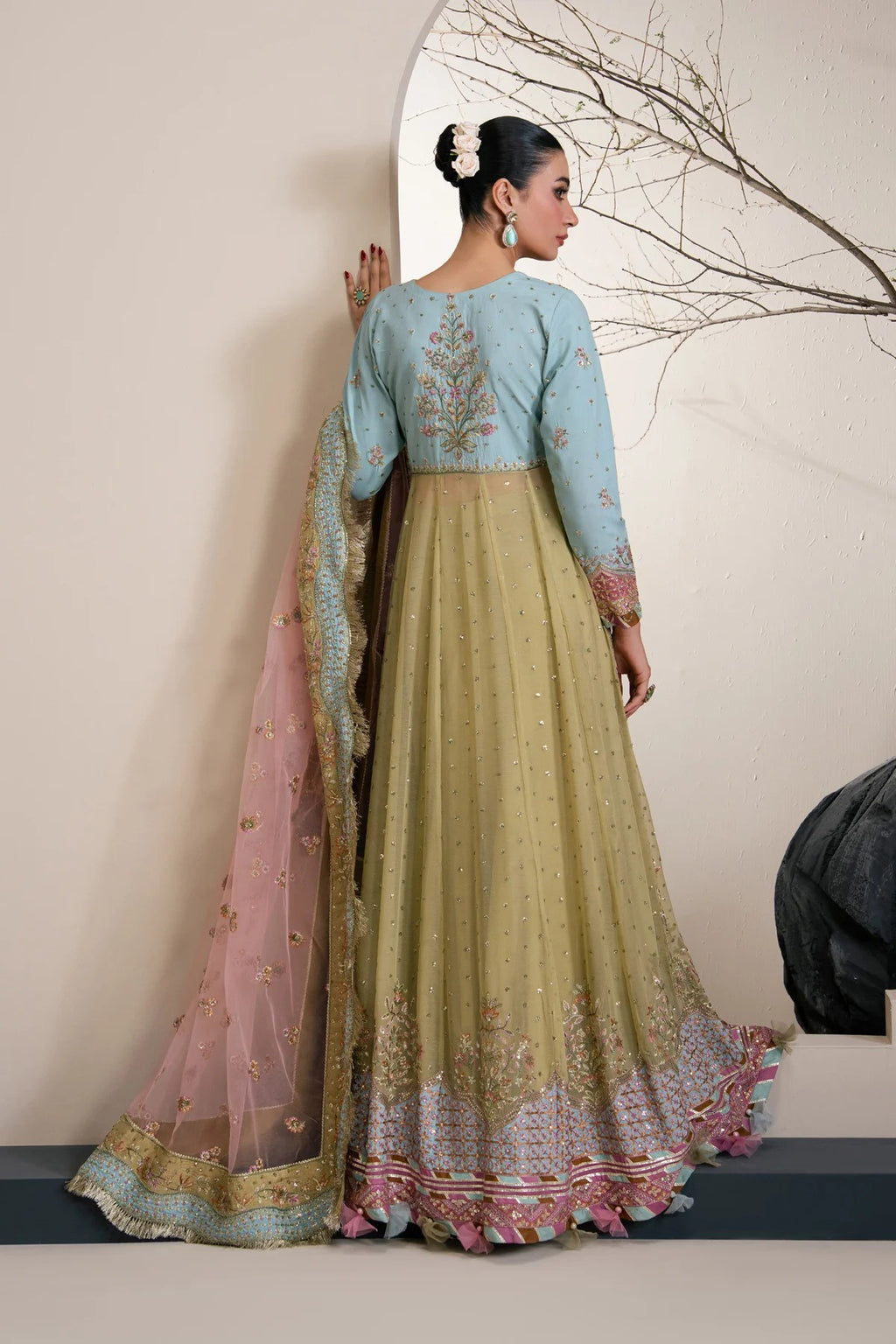 A woman modeling a traditional Indian outfit with a light blue embroidered blouse, a beige skirt with pink and blue floral embroidery, and a pink dupatta with gold detailing, posed against a neutral wall.