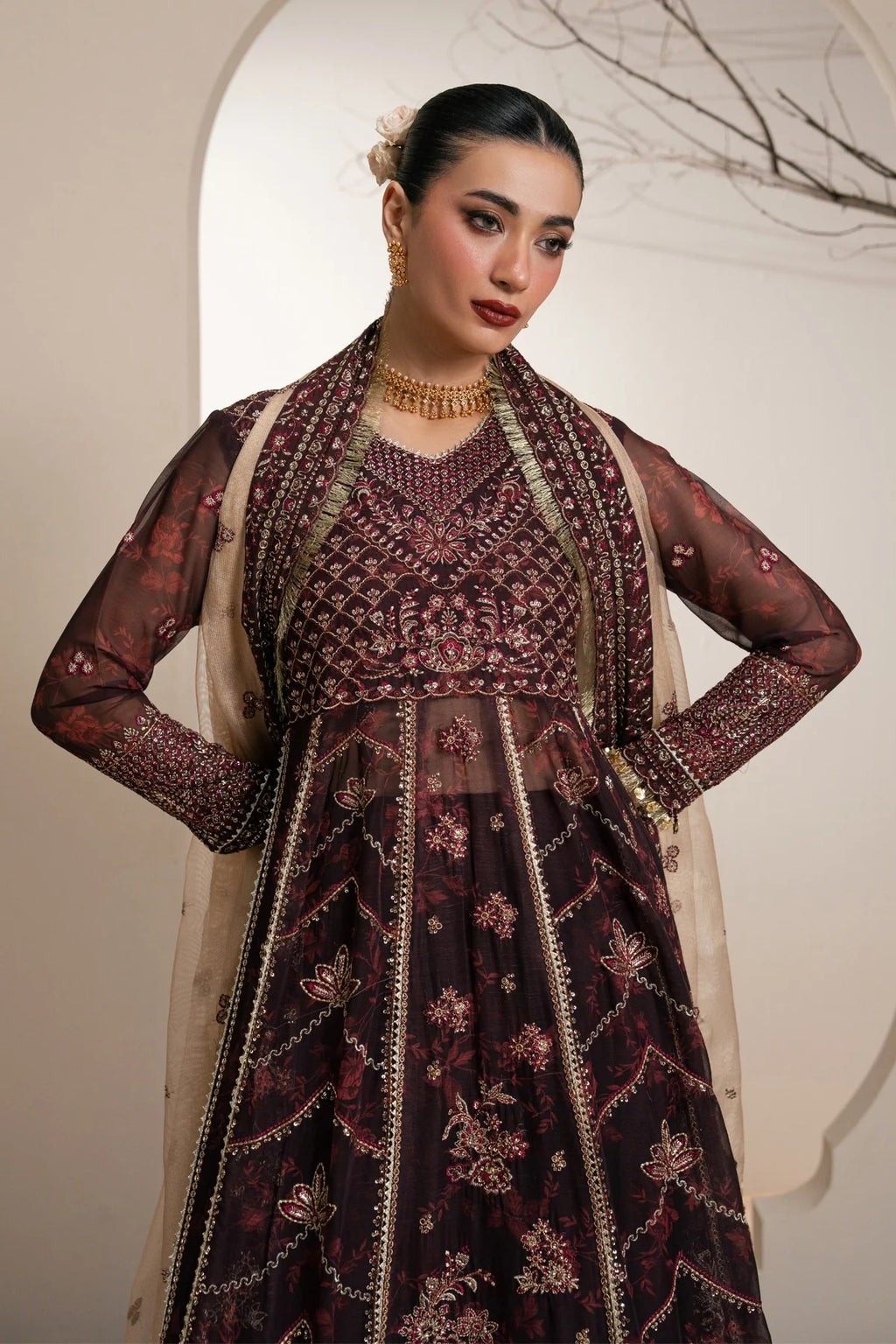 A woman models a heavily embellished dark plum purple traditional Anarkali gown with intricate gold embroidery and a beige dupatta, set against a warm beige background with an arched doorway.