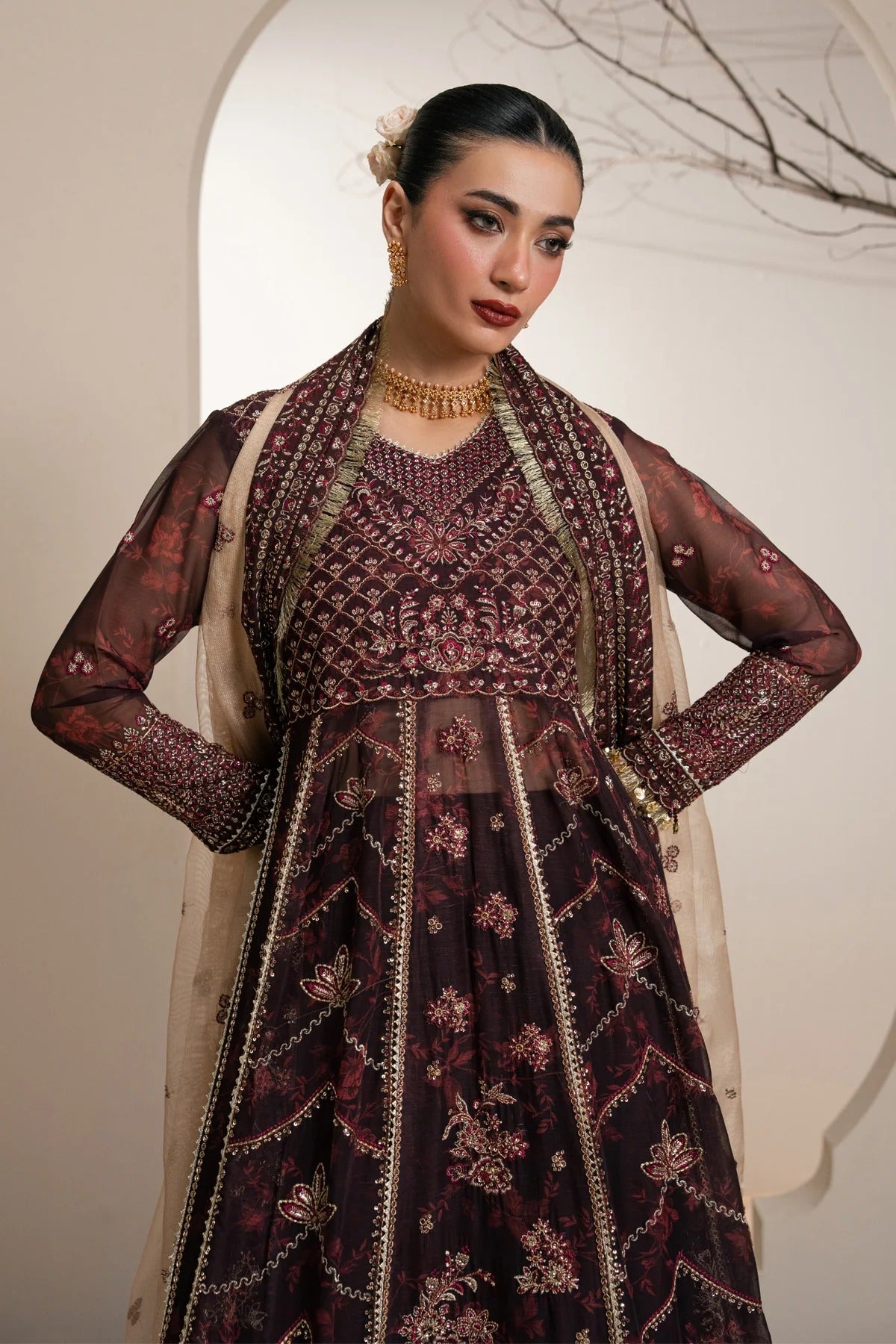 A woman models a heavily embellished dark plum purple traditional Anarkali gown with intricate gold embroidery and a beige dupatta, set against a warm beige background with an arched doorway.