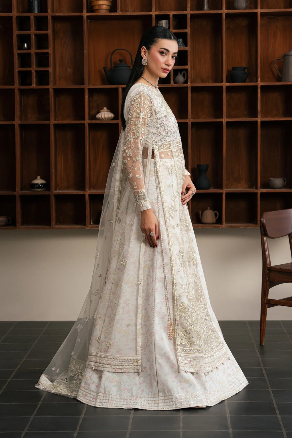 A woman models a traditional white and gold embroidered Anarkali gown with a matching sheer dupatta in a room with a wooden shelving unit in the background.