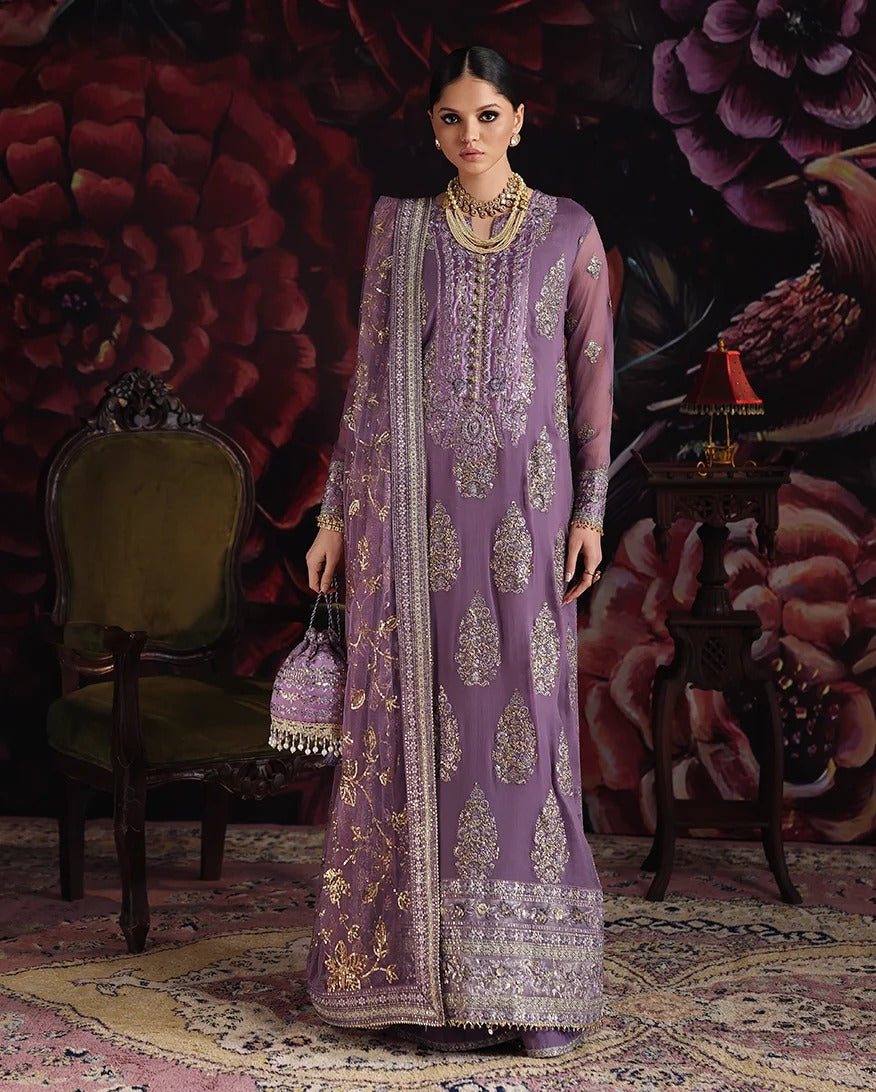 A woman models a traditional purple ethnic outfit with intricate gold embroidery, standing in a decorated room with a floral backdrop, an armchair, and a side table.