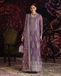 A woman models a traditional purple ethnic outfit with intricate gold embroidery, standing in a decorated room with a floral backdrop, an armchair, and a side table.