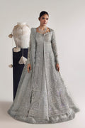 A model wears an ornate silver embellished Anarkali gown with a matching sheer dupatta, standing next to a mannequin dressed in a white embroidered outfit against a plain light gray studio background.