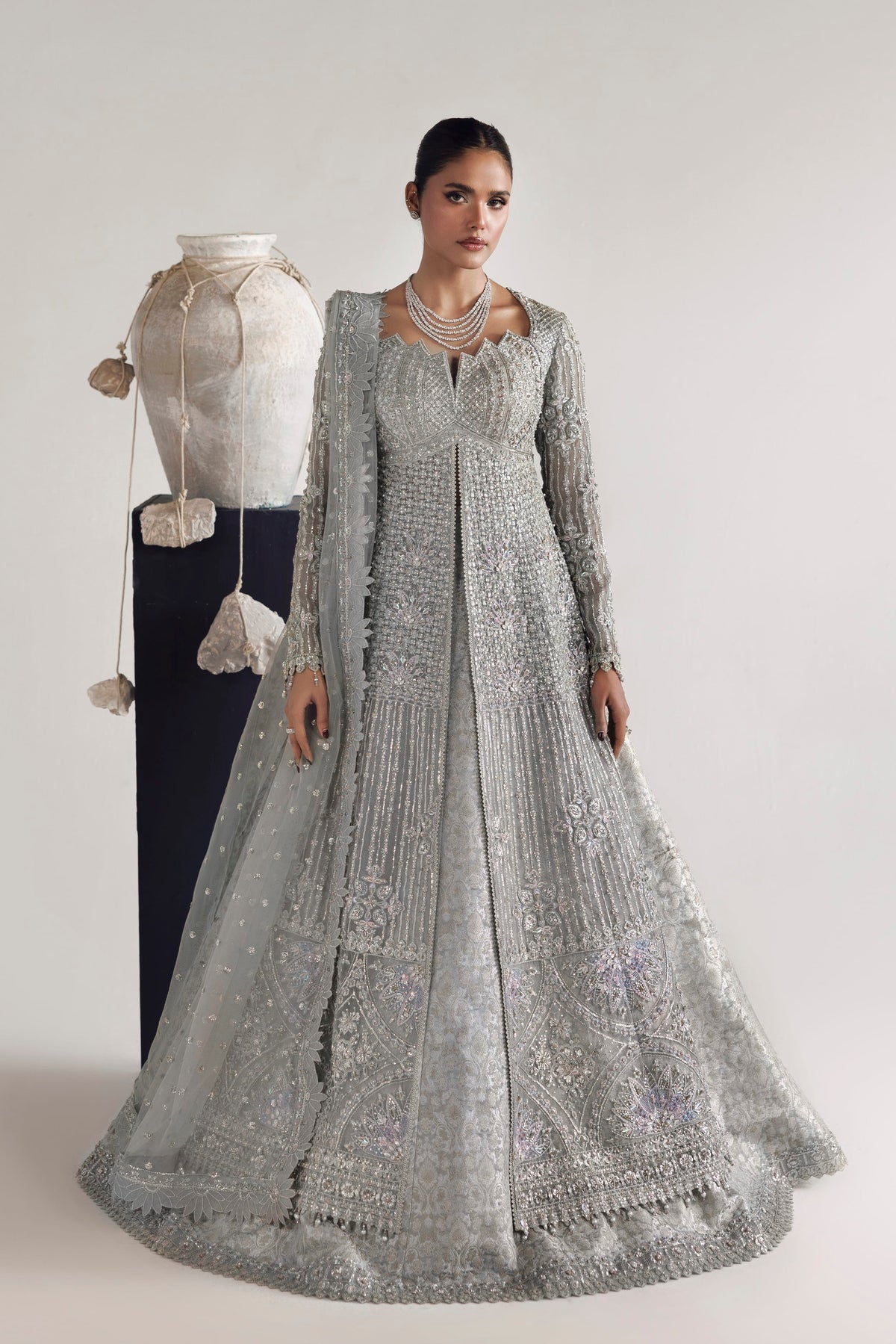 A model wears an ornate silver embellished Anarkali gown with a matching sheer dupatta, standing next to a mannequin dressed in a white embroidered outfit against a plain light gray studio background.