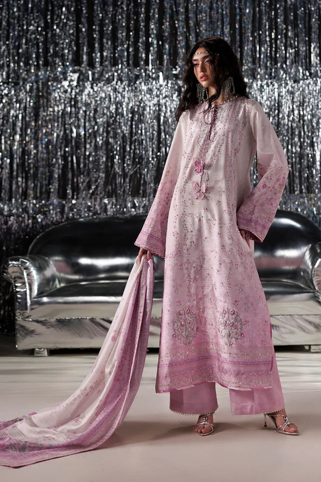 A model wearing a light pink embroidered traditional outfit with a matching dupatta, standing in front of a silver tinsel curtain backdrop.