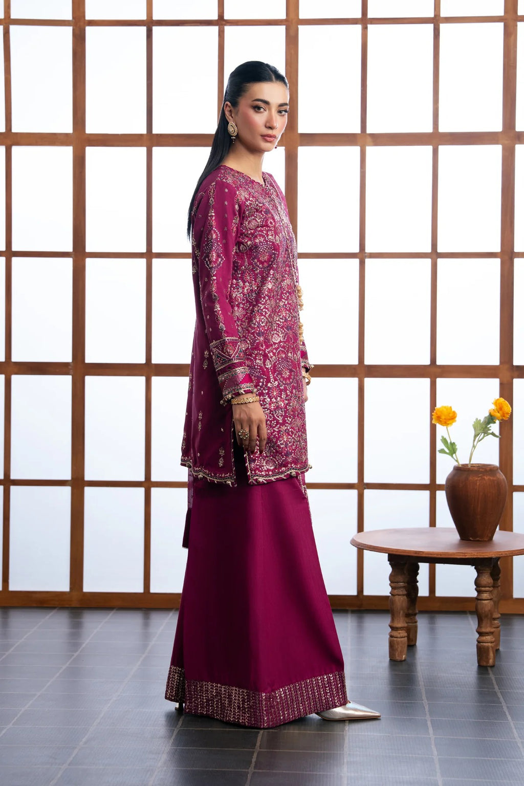 A woman models a traditional Indian outfit consisting of a magenta embroidered kurta and a matching wide-leg skirt with gold zari borders, set against a wooden lattice screen background.