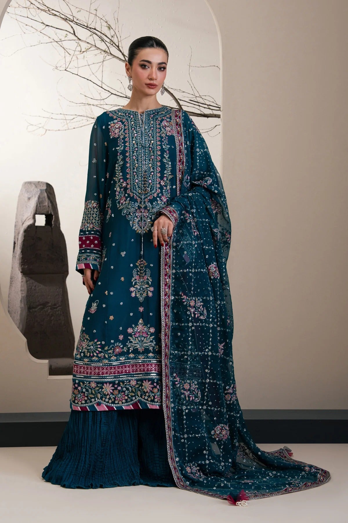 A woman models a traditional three-piece ethnic suit set in deep teal blue, featuring intricate floral embroidery, printed motifs, and a matching dupatta, set against a minimalist beige wall with a decorative branch print.