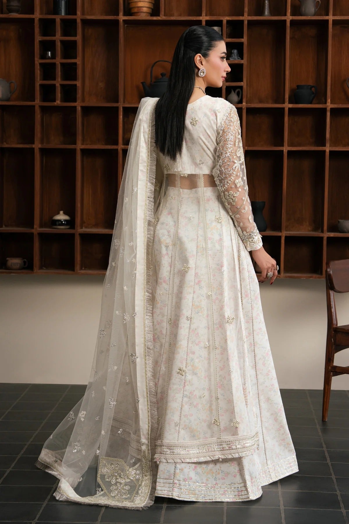 A woman modeling a traditional Indian anarkali suit from the back, featuring a cream-colored embroidered skirt, matching embroidered long sleeves, and a sheer dupatta, set against a dark wooden shelving background.