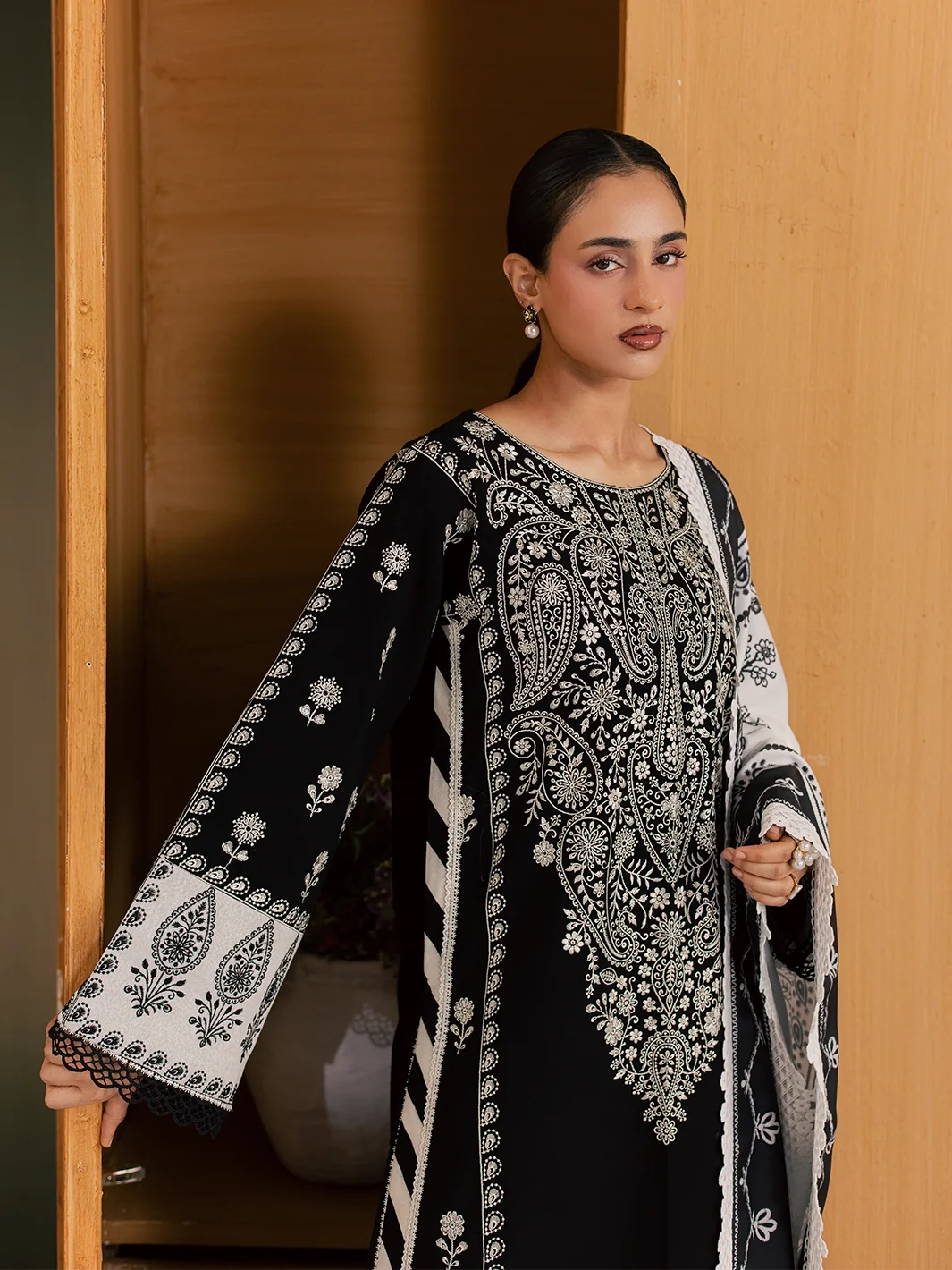 A woman models a black and white embroidered traditional outfit with a matching printed shawl, leaning against a wooden door frame.