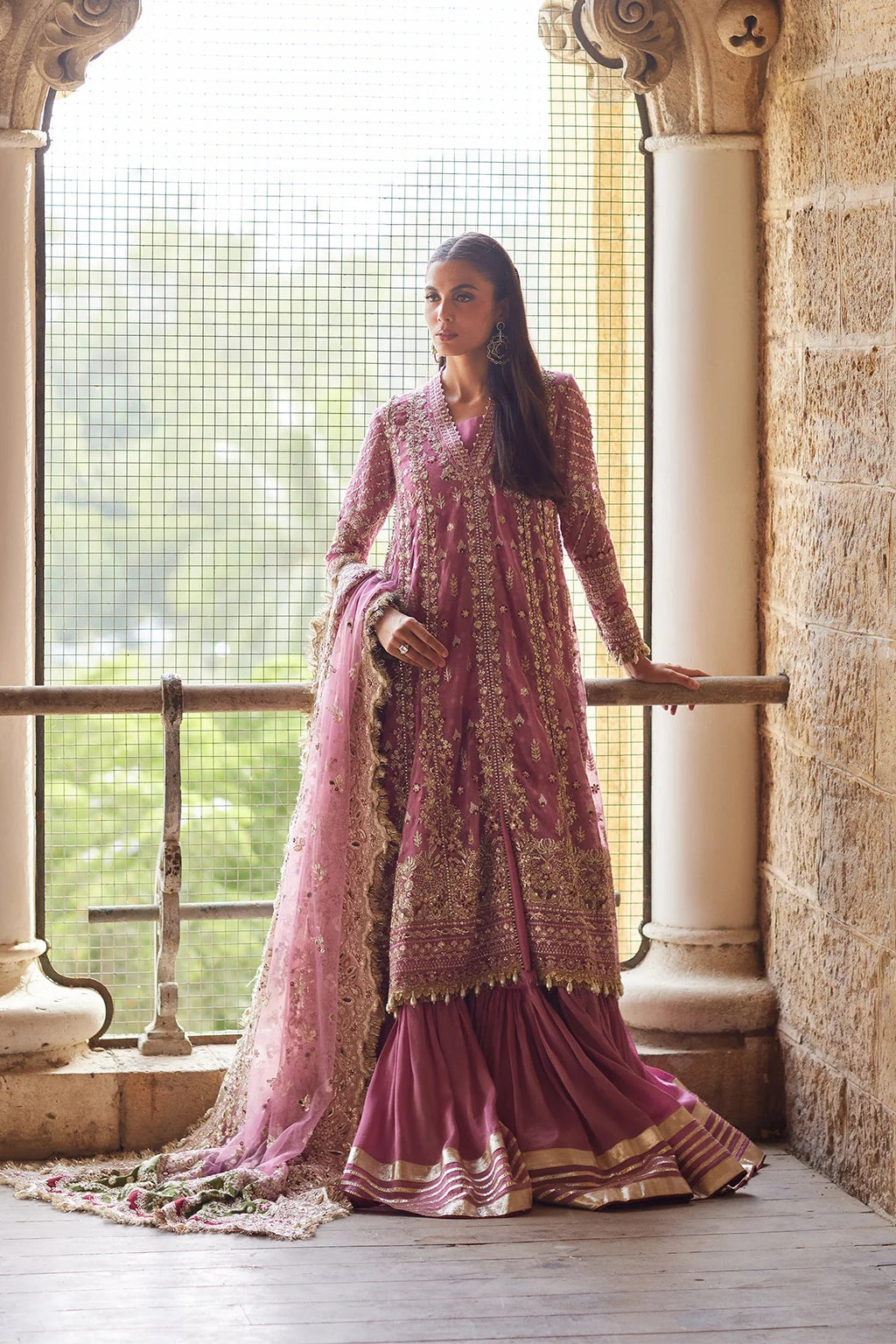 A woman models a traditional Indian salwar kameez outfit with a matching dupatta, featuring a dusty pink and gold embroidered design, set against a stone balcony backdrop.