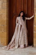 A woman models an ornate beige and gold embroidered traditional outfit with a long train, standing in front of a large wooden door and stone wall.