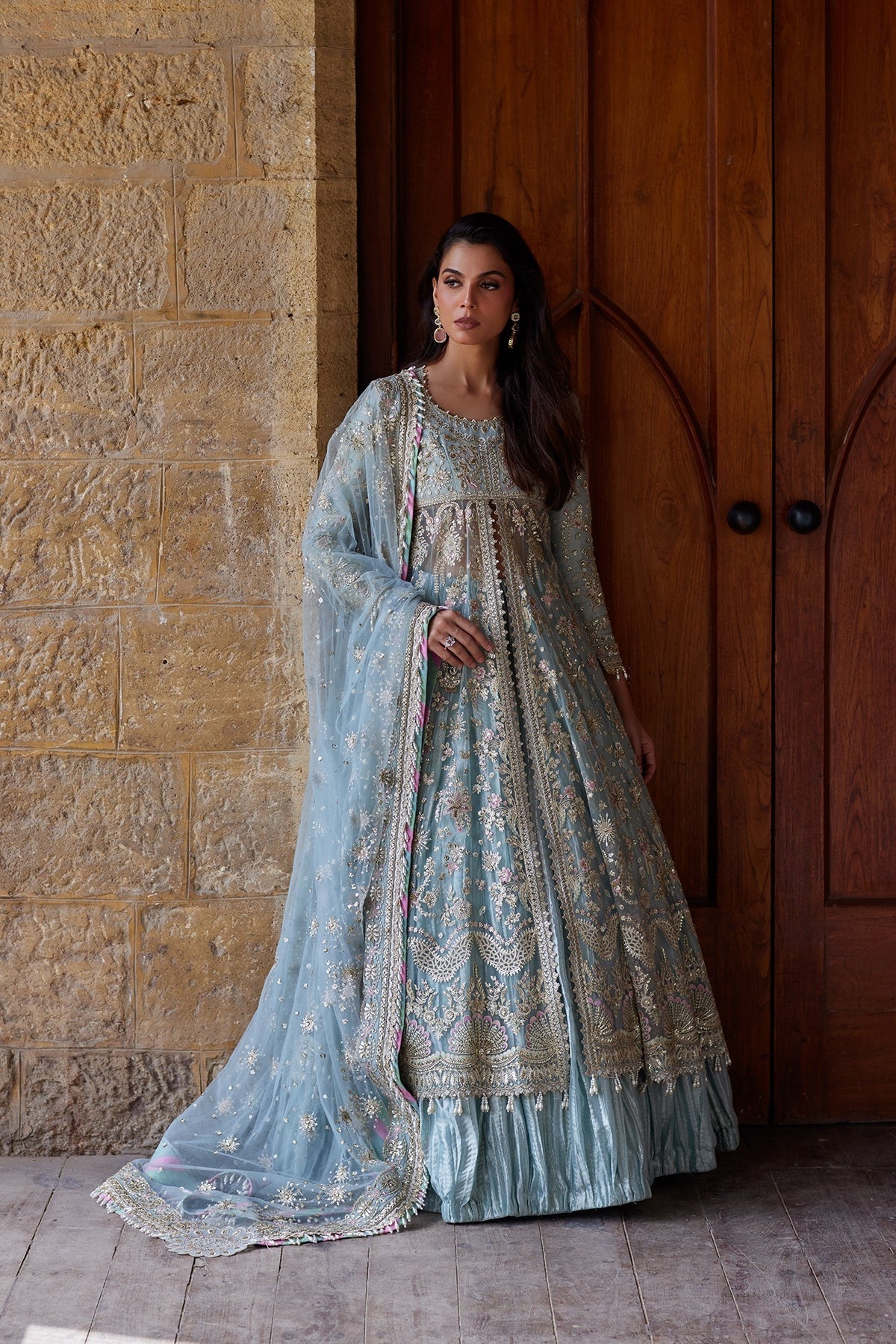 A woman stands in front of a stone wall and wooden door wearing a light blue traditional embroidered outfit with a matching sheer dupatta.