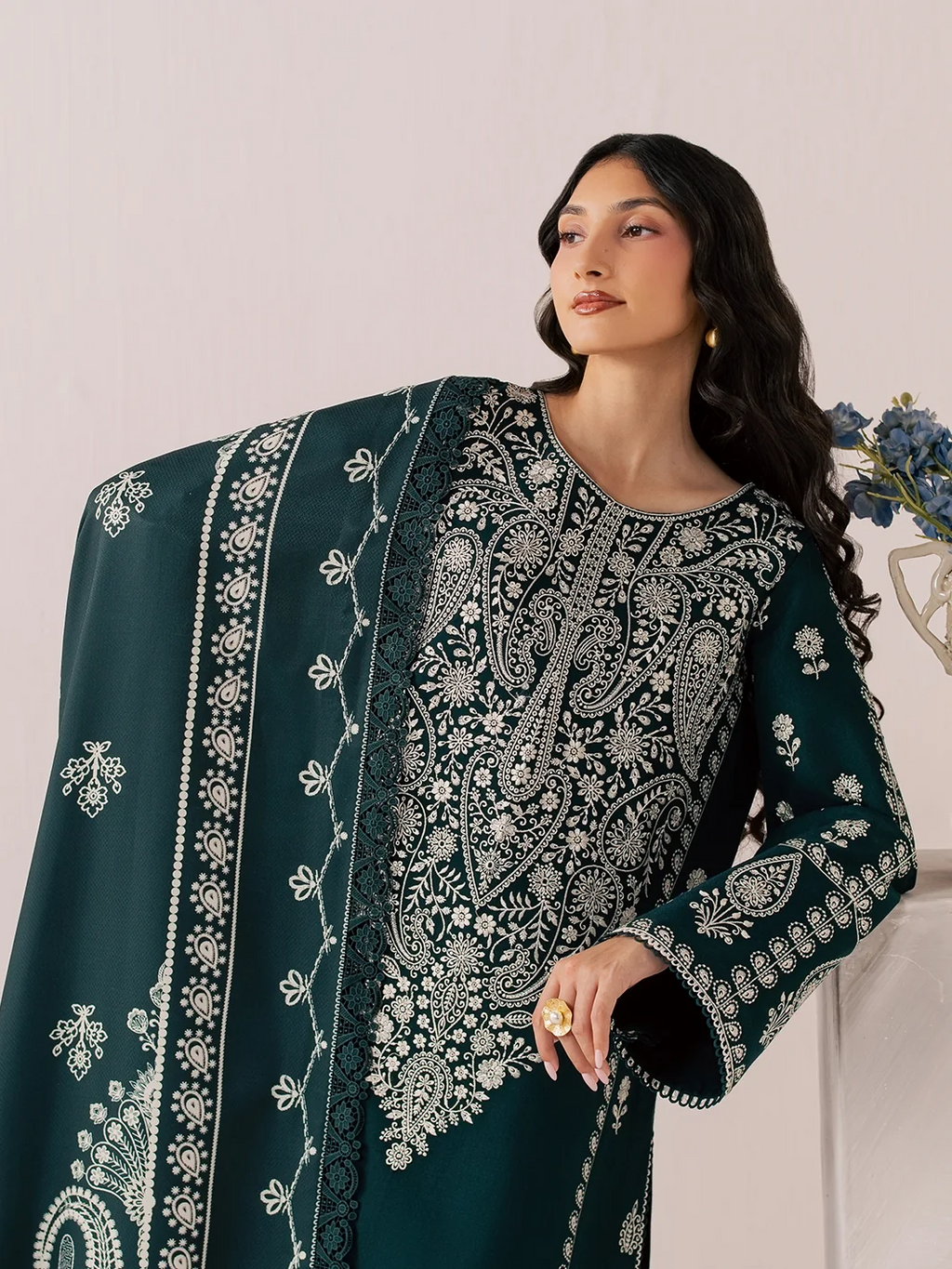 A woman models a dark teal green three-piece traditional outfit with intricate silver embroidery, matching wide-leg trousers, and a dupatta, standing in a studio setting next to a marble block with blue flowers.