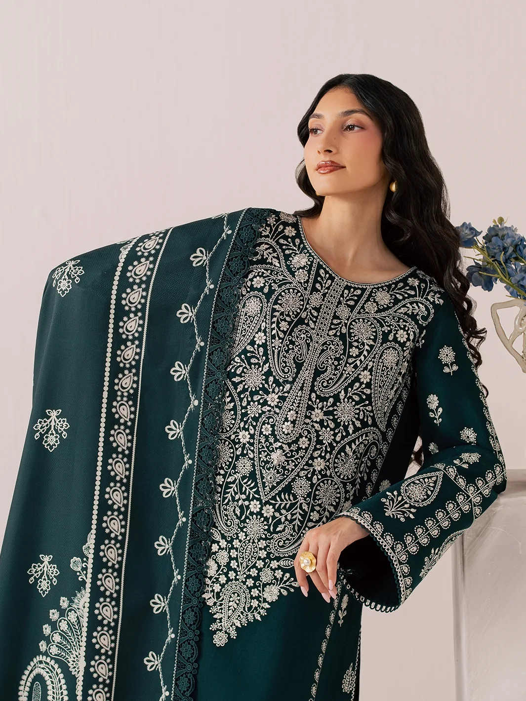 A woman models a dark teal green three-piece traditional outfit with intricate silver embroidery, matching wide-leg trousers, and a dupatta, standing in a studio setting next to a marble block with blue flowers.