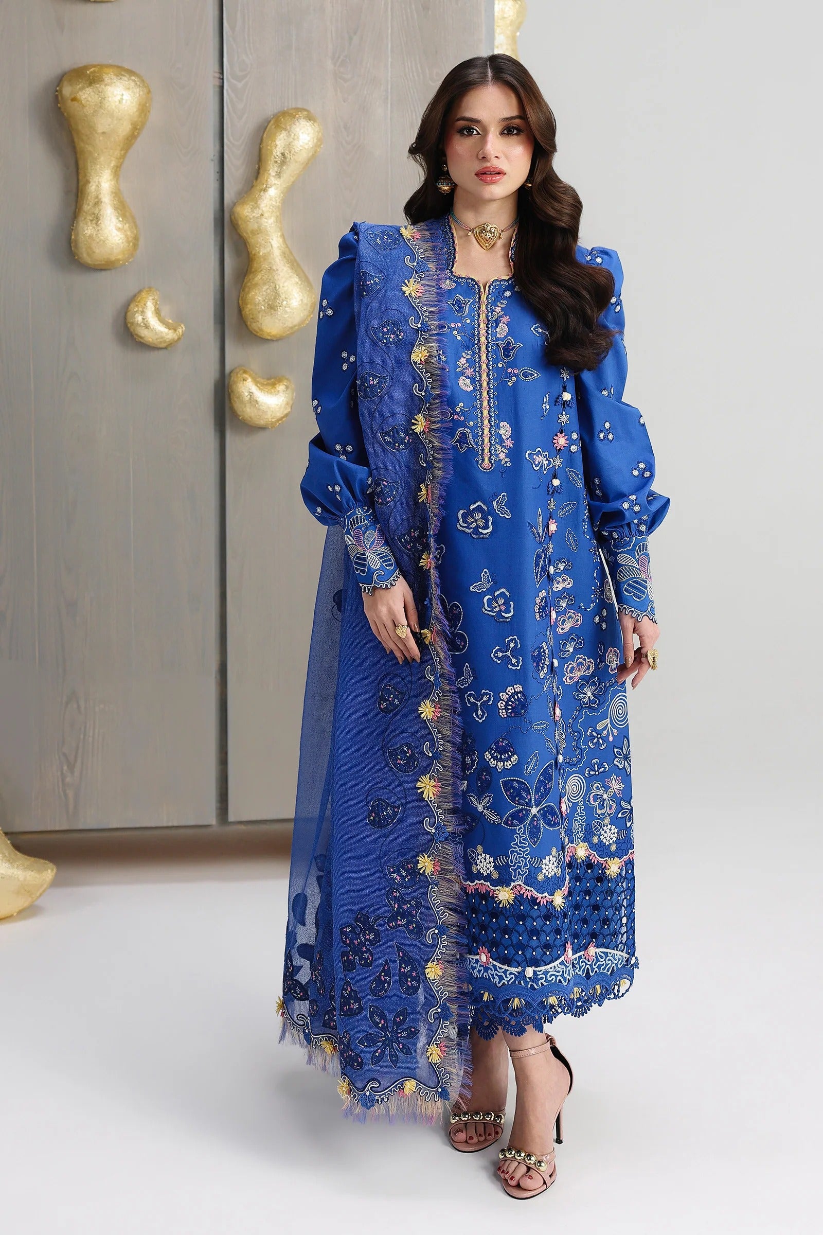 Woman wearing a blue embroidered outfit with gold details against a neutral background