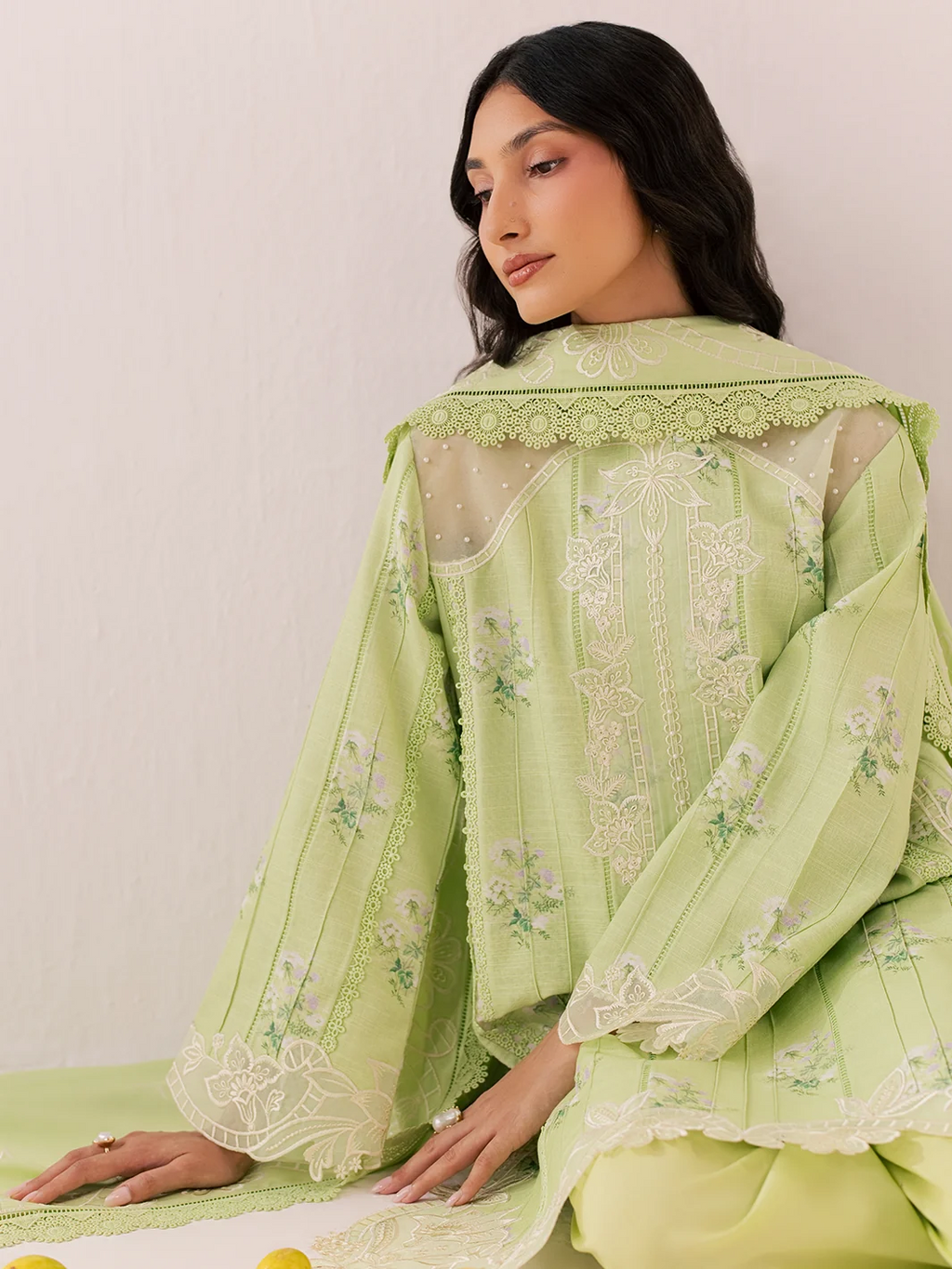 A woman wearing a light green embroidered traditional outfit with sheer sleeves and lace trim, posed against a plain light background.