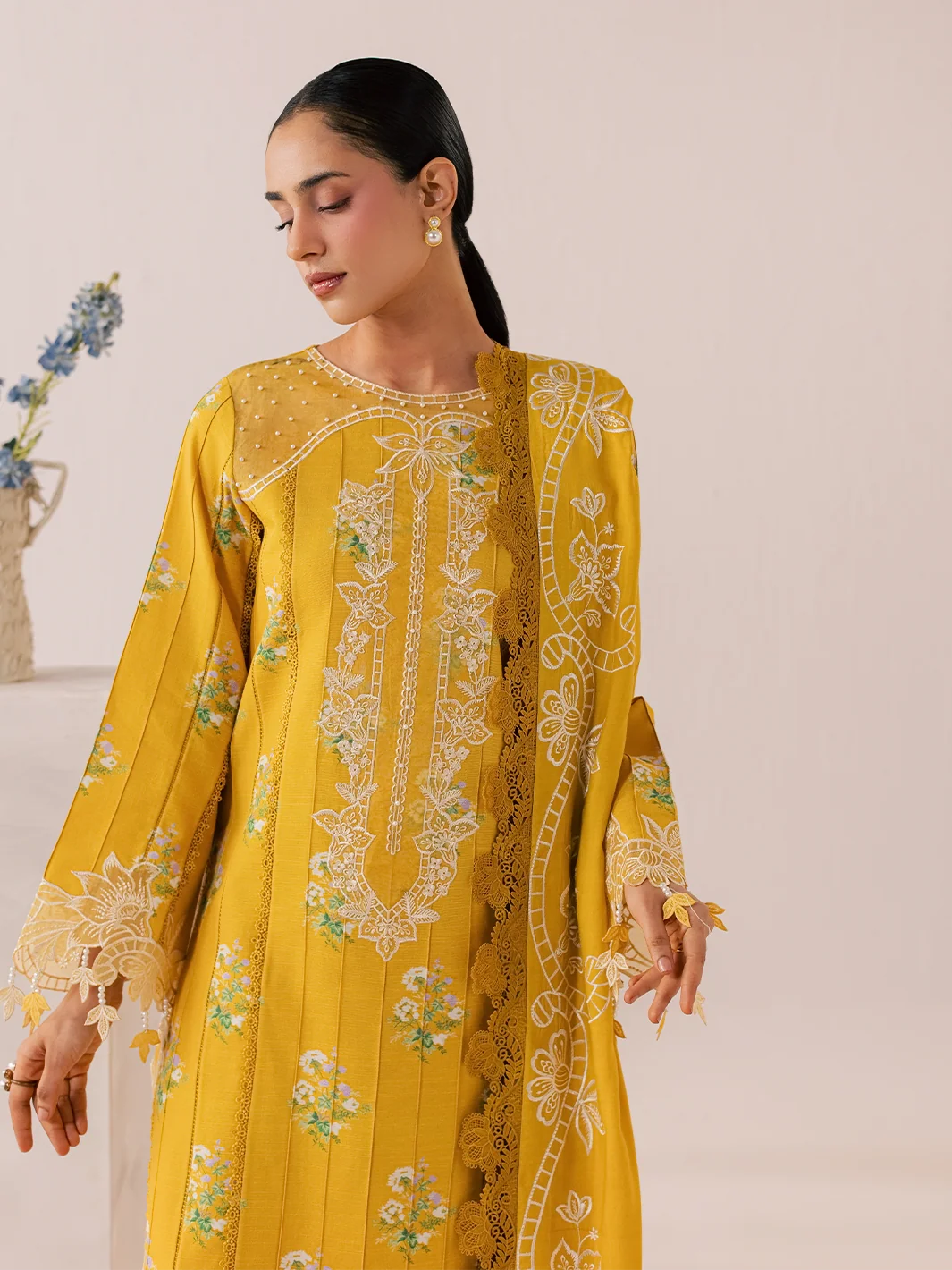 A woman wearing a bright yellow traditional embroidered outfit with white floral embroidery and lace details, photographed against a soft beige studio background.
