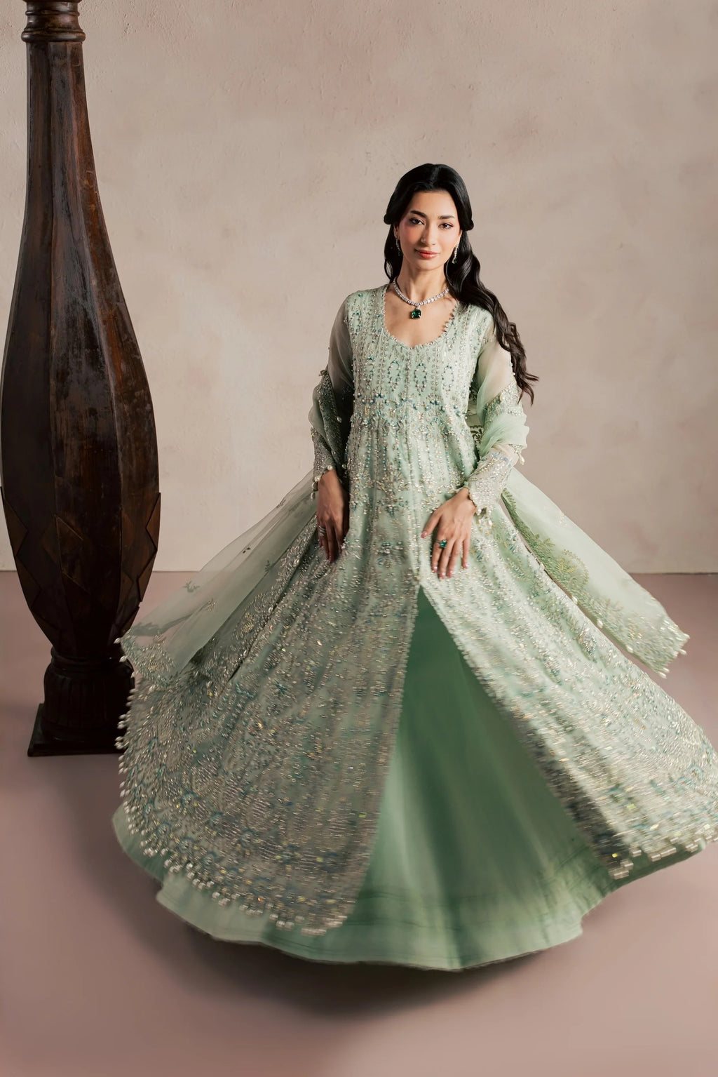 A woman models a light sage green Anarkali gown with intricate silver embroidery and sheer sleeves, standing in a studio with a textured beige wall and a dark wooden pillar.