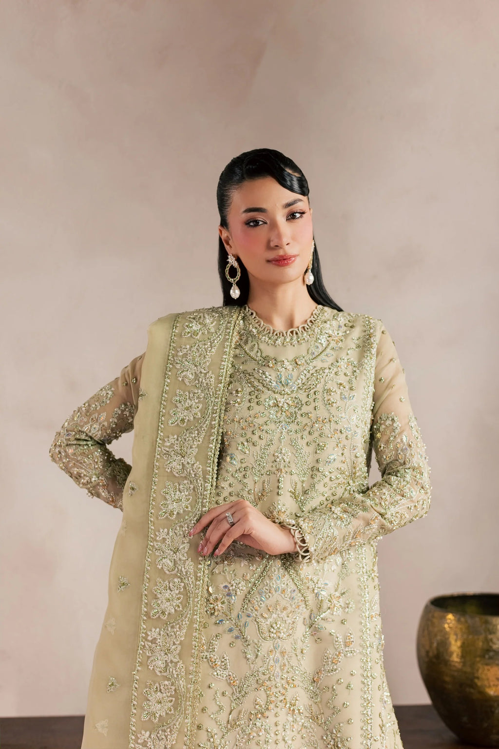 A woman modeling a traditional Indian salwar kameez suit in a light sage green color with intricate gold and silver embroidery, paired with a matching dupatta, set against a neutral beige background.