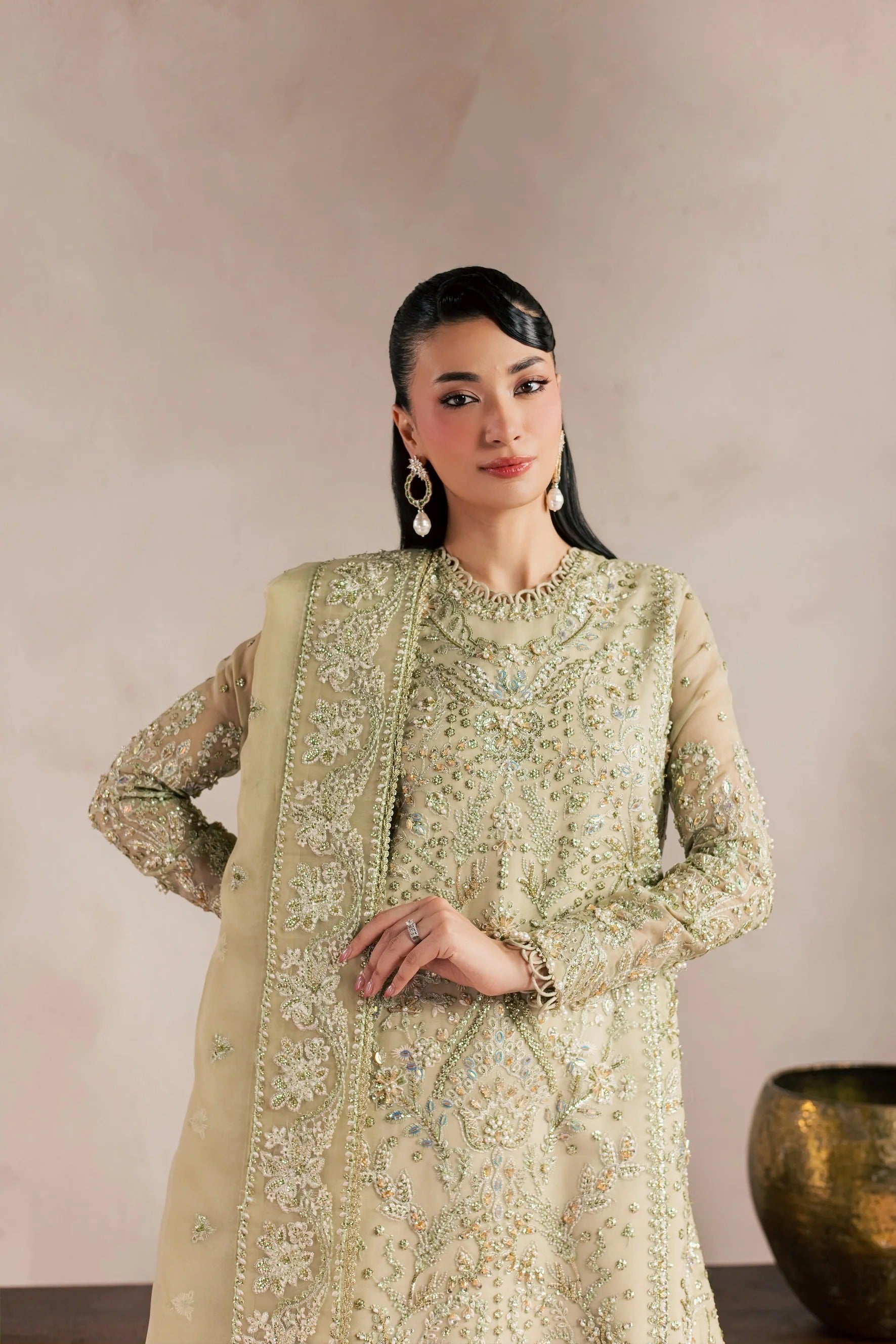 A woman modeling a traditional Indian salwar kameez suit in a light sage green color with intricate gold and silver embroidery, paired with a matching dupatta, set against a neutral beige background.