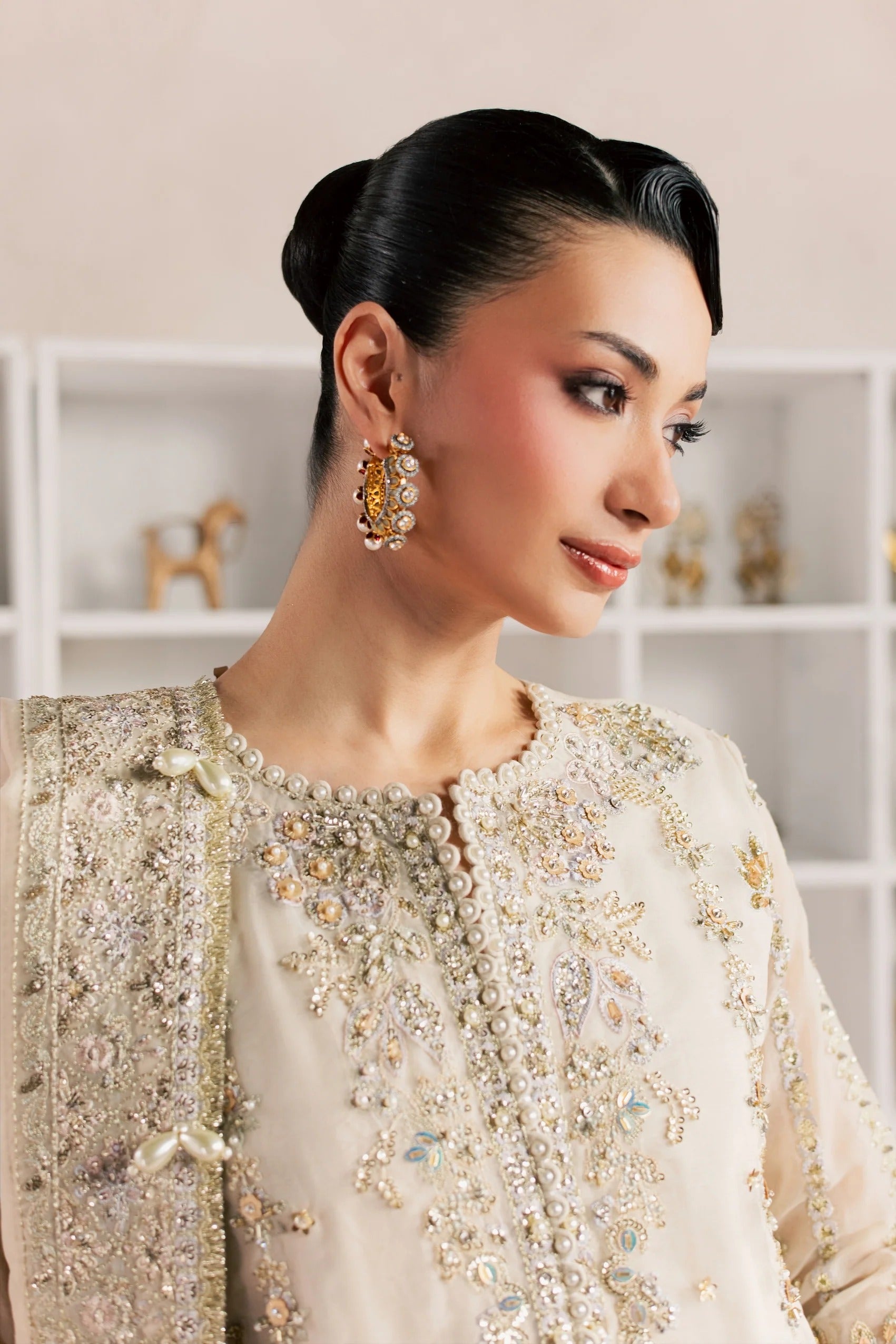 A woman wearing an ornate cream-colored embroidered traditional outfit with intricate beadwork and a matching sheer draped shawl, paired with a large gold and pearl earring, set against a soft beige background with white shelving.